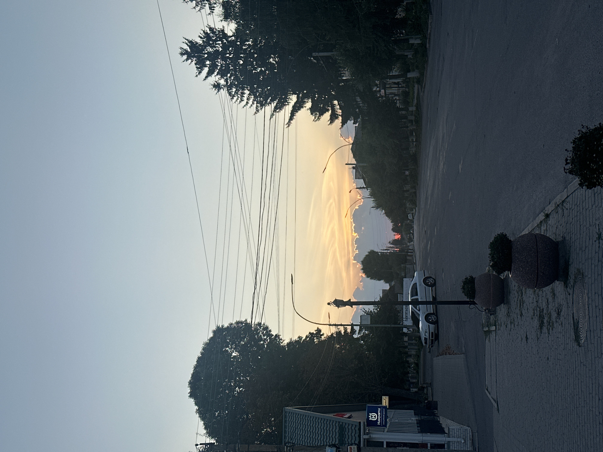 The image shows a street scene in Khmilnyk, Ukraine, at dusk. A paved road runs through the center of the frame, flanked by sidewalks and trees. The sky is a soft blue, with the setting sun visible in the distance, casting a warm glow on scattered clouds.
On the left side of the street, a building with a green roof is visible, featuring a Husqvarna sign. A white car is parked on the right side of the road. Two large planters with plants sit on the sidewalk. Overhead, power lines crisscross the sky. The overall atmosphere is calm and peaceful, capturing a quiet moment in the city.