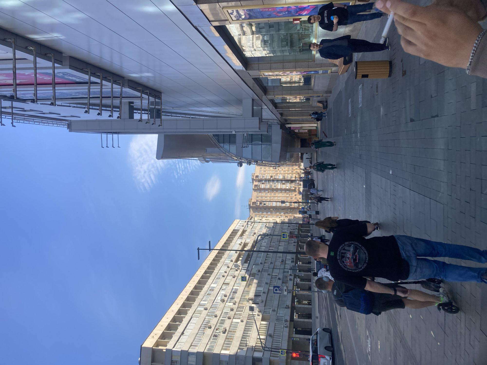 This image captures a vibrant city street scene on a bright, sunny day in Moscow, Russia. The perspective is from a pedestrian's point of view, with a person's hand holding a lit cigarette visible in the immediate foreground.
The street is lined with buildings of contrasting styles. On the left, a modern structure features a sleek, light-colored facade with extensive glass panels reflecting the sky and surrounding environment. Metal fixtures, possibly for lighting or signage, hang from its overhang. On the right, an older, multi-story residential or office building dominates, characterized by numerous windows, balconies, and external air conditioning units.
Several people populate the paved sidewalk. In the foreground, two men walk away from the camera; one wears a black t-shirt with a white circular logo on the back displaying the Russian word "ФОРСАЖ" (Forsazh), while the other pushes a kick scooter. Further down the street, two individuals in green uniforms, likely maintenance workers, are visible. Near a reflective building entrance, two men stand, one in a dark jacket holding a light blue cup, and another in a dark suit jacket. Their reflections, along with the sky and other architectural elements, are clearly visible in the glass.
The sky is a clear, brilliant blue with scattered wispy white clouds. Visible text includes "ФОРСАЖ" on a t-shirt, "4 000 ₽" (4000 Rubles) on a poster, and traffic signs like a blue "P" for parking and a red traffic light displaying the number "2". The scene depicts a typical urban environment with daily activities under pleasant weather.