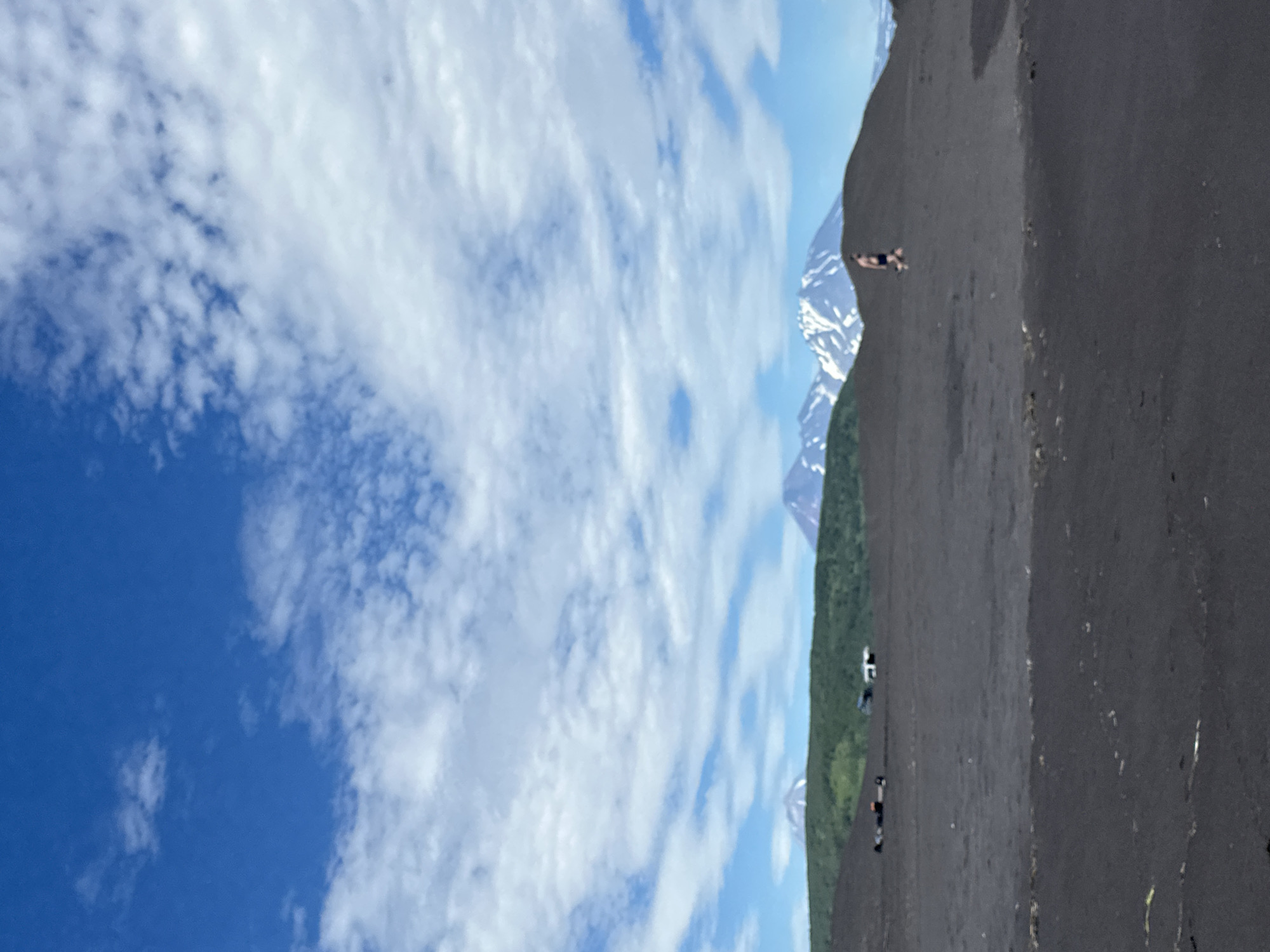 This is a wide-angle, medium-distance shot of a volcanic landscape under a bright blue sky with scattered white clouds. The foreground is dominated by dark, sandy volcanic soil, possibly a beach or barren plain. In the middle ground, green, rolling hills rise, dotted with trees. Beyond the hills, majestic snow-capped volcanoes stand in the background. The sky is a vibrant blue, with wispy white clouds that are denser towards the top of the frame. In the far distance, to the right of center, a lone figure is walking across the dark terrain. In the middle distance, to the left of center, a few dark shapes that could be vehicles are visible on the green hills. The overall impression is one of a rugged, natural environment. This scene appears to be characteristic of the volcanic landscapes found around Petropavlovsk-Kamchatsky, Russia, a city known for its proximity to active volcanoes.