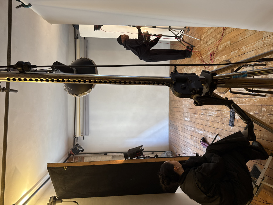 The scene depicts an active photography studio located in Paris, France. Two individuals are present amidst professional photographic equipment.
In the foreground, a person with dark hair pulled back, wearing dark clothing, is seated on a white folding stool, facing away from the camera and towards the studio's main setup. This individual has silver hoop earrings visible.
Further back and to the right, a second person, dressed in all-black attire including a dark beanie, is standing and actively adjusting a light fixture mounted on a silver tripod stand. Red electrical cables are visible coiled on the distressed wooden floorboards near this individual and extending across the floor.
Dominating the center foreground is a large, metallic studio crank stand, supporting a black light modifier (likely a softbox or beauty dish) facing towards the background. A vertical white seamless background is deployed along the far wall of the studio. To the left, a large black V-flat or reflector panel stands vertically, partially obscuring an exposed brick wall section. Additional studio lights and seamless paper rolls (one white, one grey) are mounted high on the far wall. The room features white painted walls, a high ceiling with exposed piping, and a small fan visible on the left near the floor. A white vertical panel stands on the extreme right, partially out of frame. The overall environment suggests a state of preparation or active operation for a photoshoot.