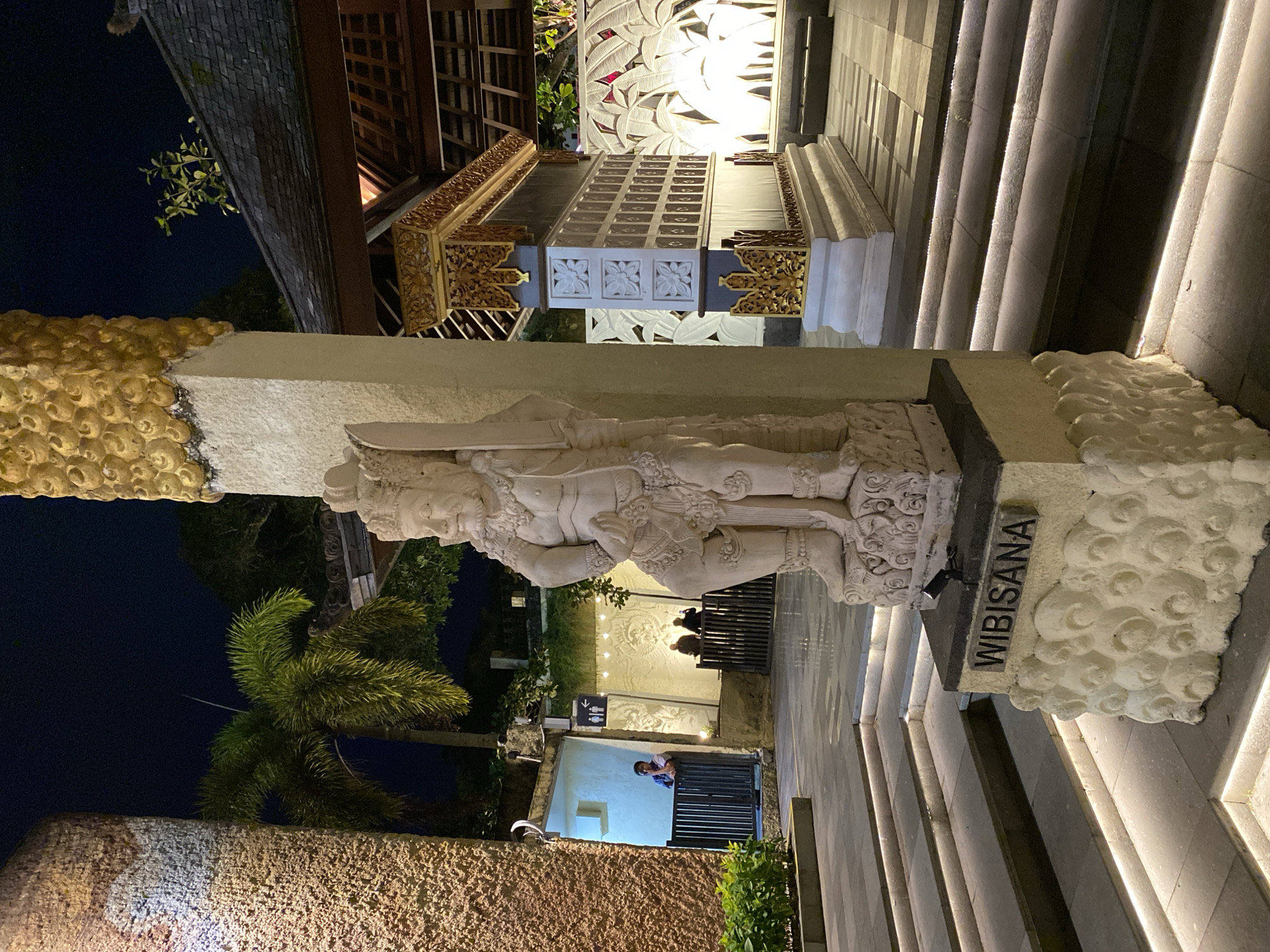 In Kuta Selatan, Indonesia, a nighttime scene unfolds at what appears to be a temple or cultural complex. A prominent stone statue of a figure, identified by the name "WIBISANA" inscribed on its base, stands in the foreground. The statue is intricately carved and weathered, with detailed facial features and ornate clothing. It is positioned on a decorative pedestal adorned with a repeating circular pattern.
To the right of the statue, a section of a building with a dark tiled roof is visible. This structure features ornate golden carvings along its roofline and decorative elements on its walls, including a section with square panels. The building is illuminated by interior lights that cast a warm glow. Beyond this structure, a wall with a pattern of large, stylized leaves is illuminated by white lights, creating a dramatic backdrop.
To the left of the statue, a textured, tan-colored cylindrical structure or pillar is partially visible. Further in the background, there is a palm tree with lush green fronds silhouetted against the dark night sky. The ground is paved with large square tiles, and a set of stone stairs with integrated strip lighting leads up from the foreground, creating illuminated pathways.
In the middle ground, to the left, a section of an open-air corridor or walkway is visible. A person, appearing to be a man wearing a blue shirt, leans against a dark metal railing, looking into the distance. This area is brightly lit, revealing white walls and a sign with pictograms of a male and female figure, indicating restrooms. Further to the right, in a dimly lit area, two individuals are seated behind another dark metal railing. The overall ambiance is one of cultural display and nighttime illumination, suggesting a public or tourist-oriented space.