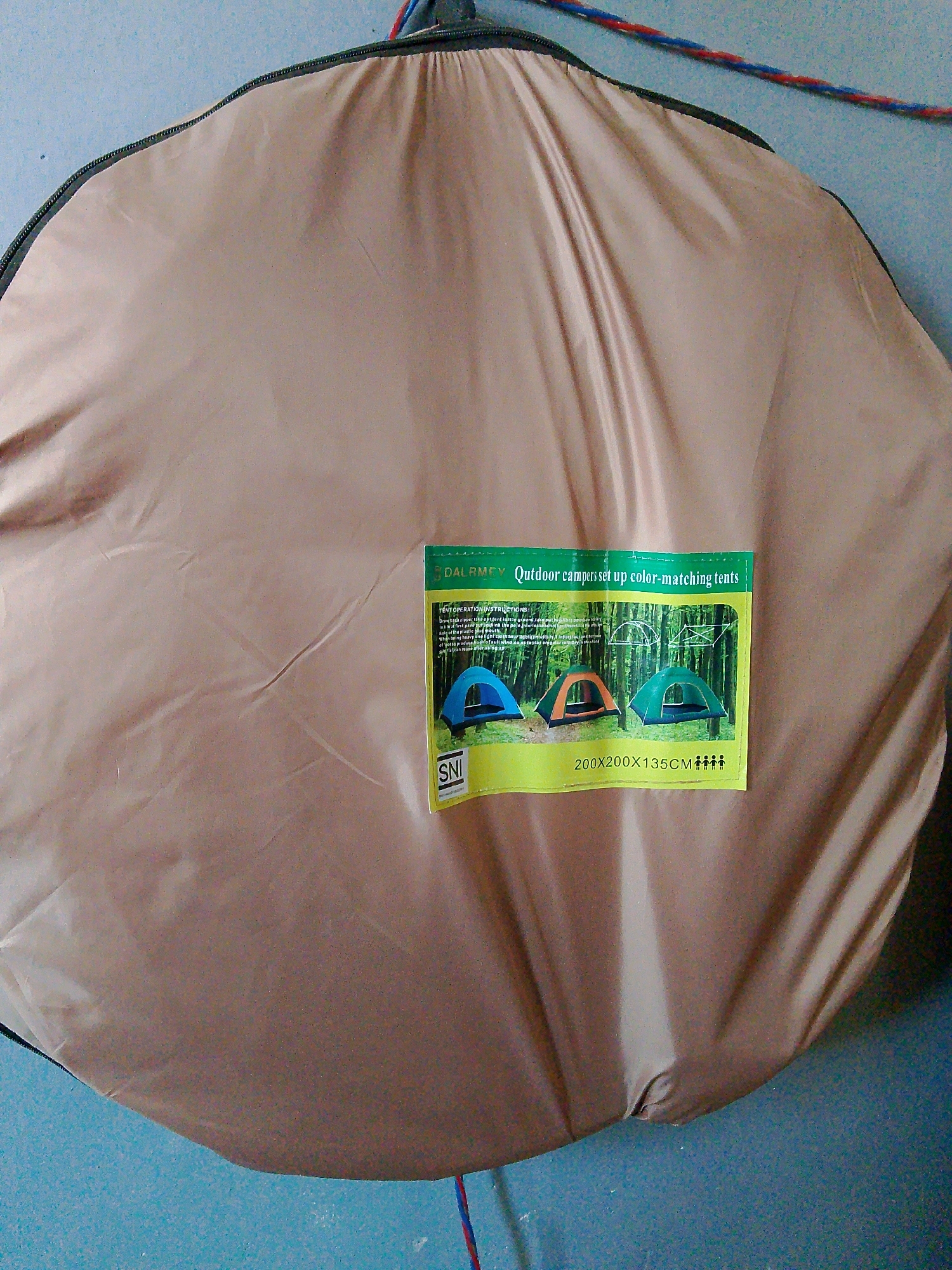 A beige tent bag is hung against a light blue wall. The bag is round and appears to be made of a lightweight, synthetic material. It is closed with a black zipper that runs along the outer edge. A small, rectangular label is attached to the front of the bag. The label is yellow and green and features text and images of tents. The text on the label reads "DAL RMEY Outdoor campers set up color-matching tents" and "SNI 200x200x135CM". There are also illustrations depicting how to set up a tent. A red and blue cord is visible hanging from the top of the tent bag. The floor is a dark blue. The scene is indoors, as suggested by the wall and the manner in which the bag is hung. It is not possible to determine the specific location within the city of Kuripan Utara, Indonesia from the image alone.
