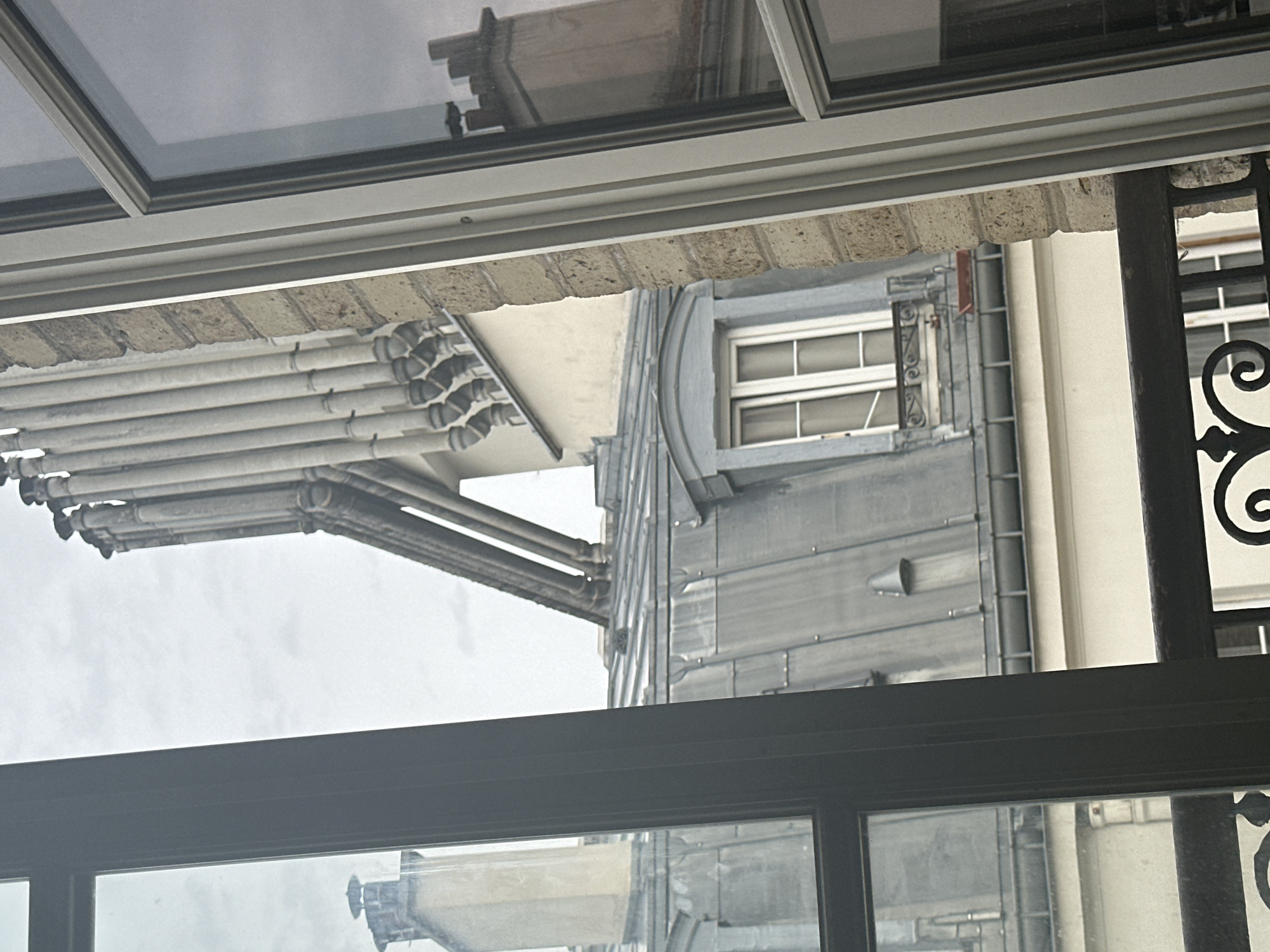 An urban scene is depicted through a window, showcasing Parisian rooftops and architectural elements. The view is framed by the dark mullions of a window on the left and the lighter frame of another window on the right, with a brick pillar separating the two perspectives.
The dominant elements in the composition are the distinctive grey, mansard-style roofs, characteristic of Parisian architecture. Multiple chimneys are visible, some grouped together in a row, others standing individually. One prominent feature is a dormer window with a blue-grey frame, set into a sloped roof, featuring multiple panes of glass and decorative ironwork below. Another dormer window, also with a decorative iron element, is visible further down.
A series of large, vertical pipes, likely for drainage or ventilation, ascend along the side of a building, adding a utilitarian detail to the urban landscape. The buildings appear to be constructed from light-colored stone or stucco, with brickwork visible in the section between the windows.
The sky is overcast and light grey, suggesting a cloudy day. The overall impression is one of a dense, historic urban environment. There are no people or animals visible in the image. The image is a still shot with no discernable action taking place.