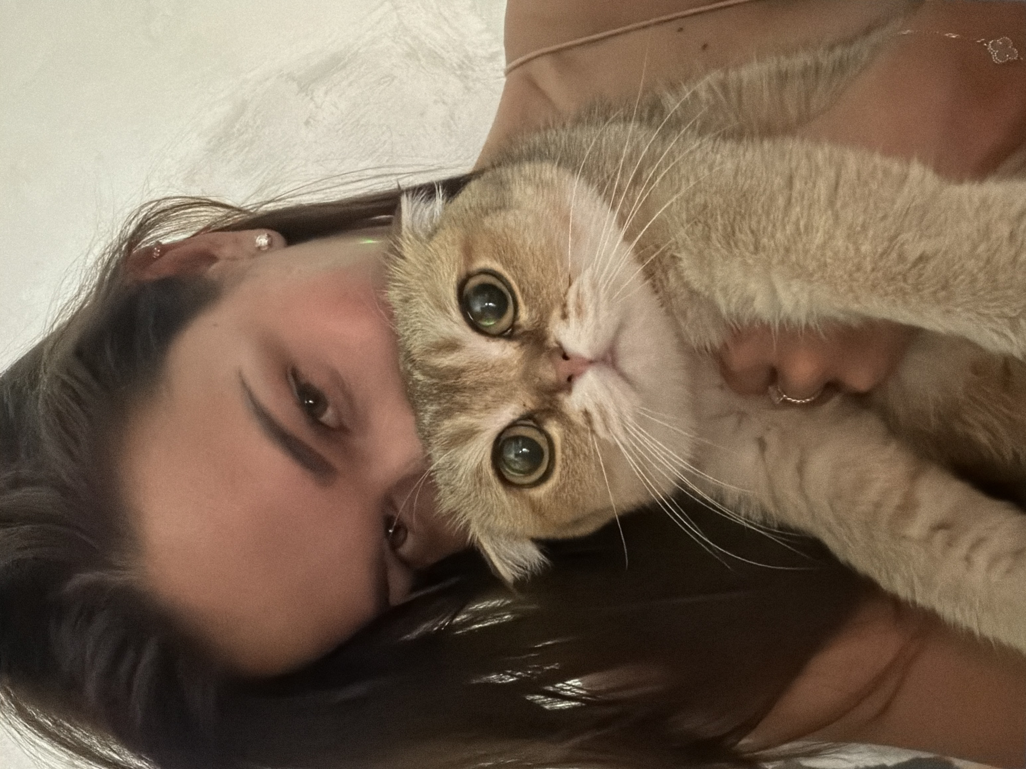 A close-up, selfie-style portrait shows a young woman holding a Scottish Fold cat. The woman has dark hair, brown eyes, and is wearing a thin strap top. She is holding the cat, which has golden-brown fur and large, round eyes, in her arms. The background is a textured, light-colored wall. There is no discernible indication of the city of Khimki, Russia, or any specific urban environment.