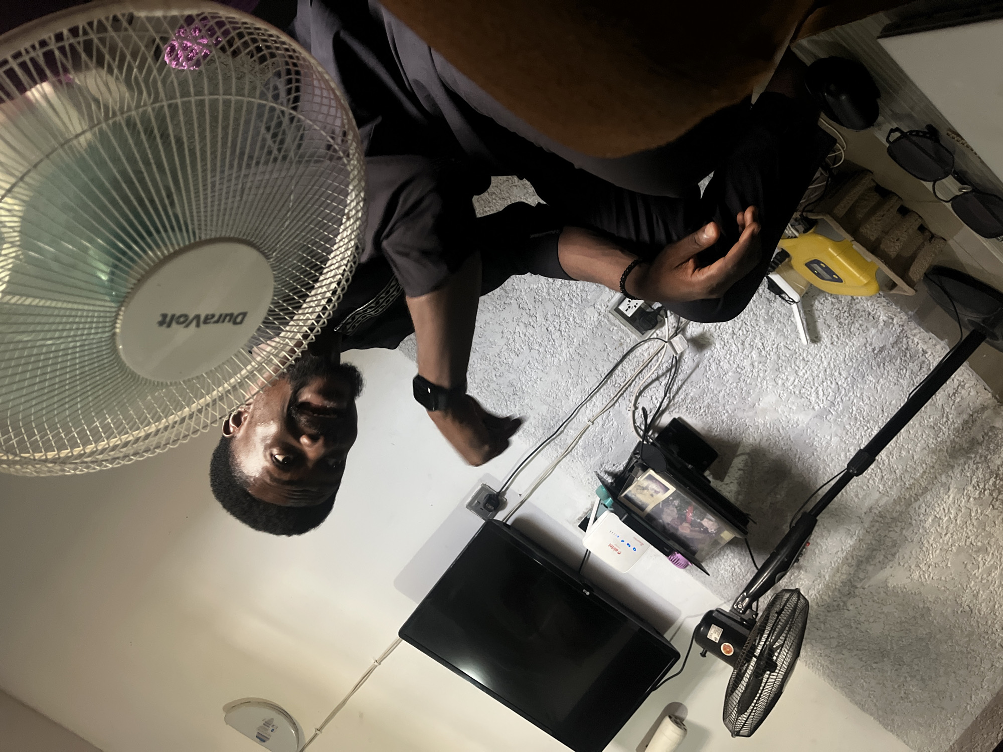 The image depicts an indoor scene within a domestic setting in the city of Abuja, Nigeria. One adult male, appearing to be of African descent with dark skin, a short haircut, and a beard, is prominently featured on the right side of the frame. He is looking upwards and to his right, with his mouth open in an expression suggestive of a smile or laughter. He wears a dark-colored, long-sleeved garment, possibly a tunic, with discernible patterned trim around the neckline. A black wristband or smartwatch is visible on his left wrist, and a black beaded bracelet is on his right wrist.
In the immediate foreground, obscuring part of the man's lower face and body, is the white-grilled head of a "DuraVolt" brand standing fan, which appears to be operational given the blurred fan blades. A purple hue or object is faintly visible within the fan's grille at the bottom.
The background reveals a textured, light-colored or white wall. A black LG flat-screen television is mounted on this wall. Below the television, a small shelf holds various electronic devices, including a white "Airtel" branded Wi-Fi router or modem with multiple indicator lights, and a stack of black electronic components. Several small framed pictures or objects are also present on this shelf. Multiple power cables and data lines are visible, extending from the electronic devices and the television to a white wall outlet located beneath the TV.
On the floor or a low platform in the lower-left quadrant, a black standing fan is positioned. Further right on the floor, a yellow power strip or adapter, a pair of black sunglasses, a black computer mouse, and a portion of what appears to be a laptop or tablet are discernible. In the upper-left, a white cylindrical object rests on a ledge, and in the upper-right, a white circular fixture is mounted on the ceiling. The overall lighting is bright, illuminating the scene.