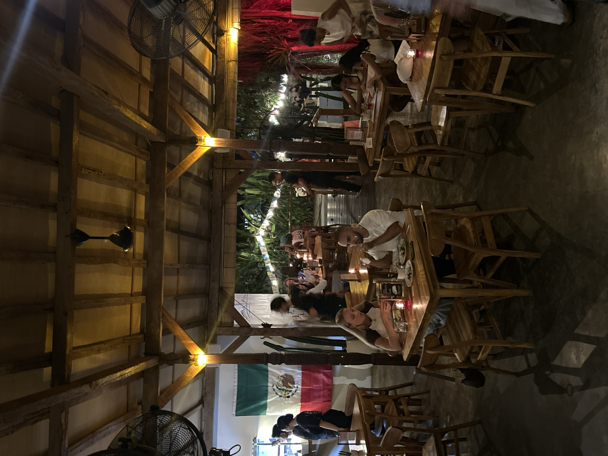 This is a medium shot, taken at night, of an outdoor restaurant with a thatched roof supported by wooden beams. The restaurant appears to be located in Kuta Selatan, Indonesia, given the presence of Mexican flags, possibly indicating a theme. A large Mexican flag hangs on the left side of the image, draped over a white wall. The restaurant is bustling with activity. Several tables are occupied by patrons eating and conversing. In the foreground, a couple is seated at a table, with the woman looking down at something on the table and the man eating. To the right, another table is occupied by two young women who are looking at menus. Further in the background, more diners are visible, as well as staff members circulating. The lighting is dim, with overhead lights and candles on the tables creating a warm, intimate atmosphere. The floor is concrete and reflects the light from the tables. Two large industrial fans are visible on the left and right sides of the image, suggesting a warm climate. Lush green foliage is visible in the background, indicating an outdoor setting.