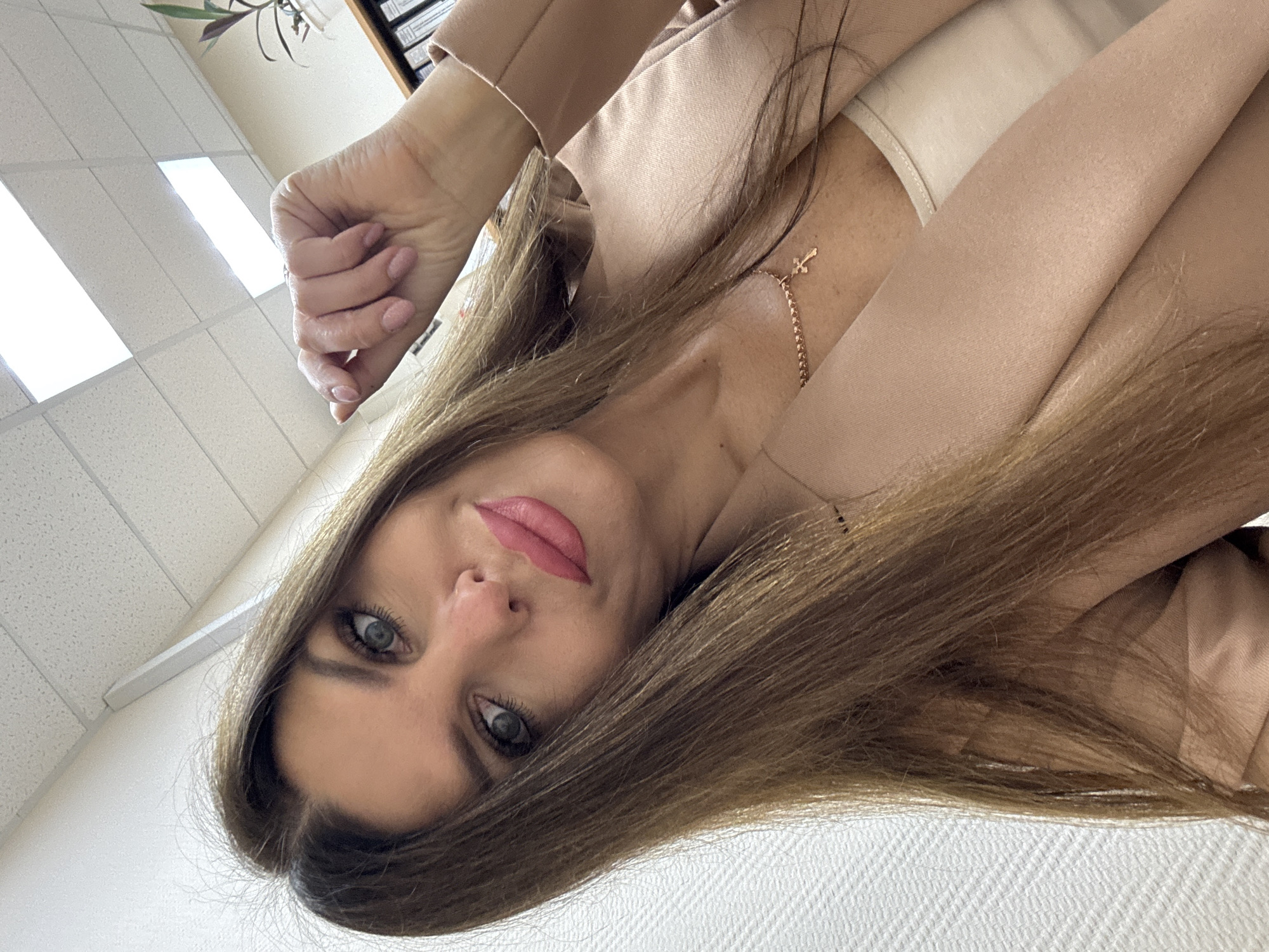 A woman is taking a selfie indoors. She has long, brown hair and is wearing a beige blazer over a light-colored top. A gold necklace with a cross pendant is visible. Her right hand is raised near her head. The background shows a textured white wall, a drop ceiling with fluorescent lighting, and a bookshelf with files and a potted plant. The image is cropped to focus on the woman's face and upper body. There is no discernible information that suggests a specific location within the city of Chaikovsky, Russia.