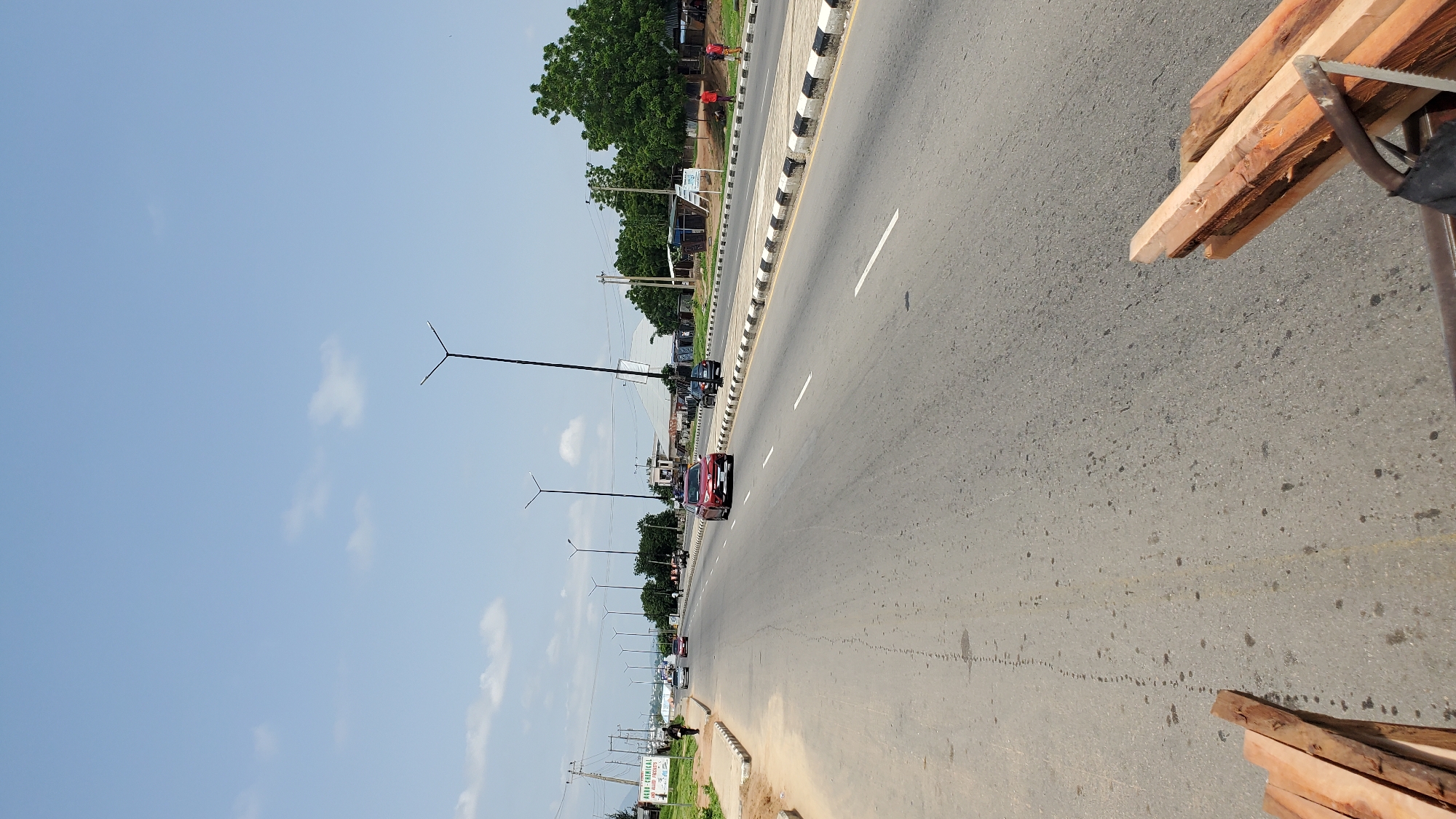 This is a street scene in Jalingo, Nigeria. A wide asphalt road with white dividing lines stretches into the distance. On the left side of the road, a sandy shoulder leads to a sidewalk with a curb. A tall street lamp with a Y-shaped head stands near the sidewalk. Further down the road, a few vehicles are visible, including a red car in the right lane. The right side of the road features a similar sidewalk and curb, also lined with street lamps. Beyond the sidewalk, there are buildings, trees, and more street lamps. In the foreground of the image, a pile of rough-cut lumber is visible, positioned to the right of the frame, suggesting it is on a vehicle or being transported. The sky is a clear blue with a few scattered white clouds.