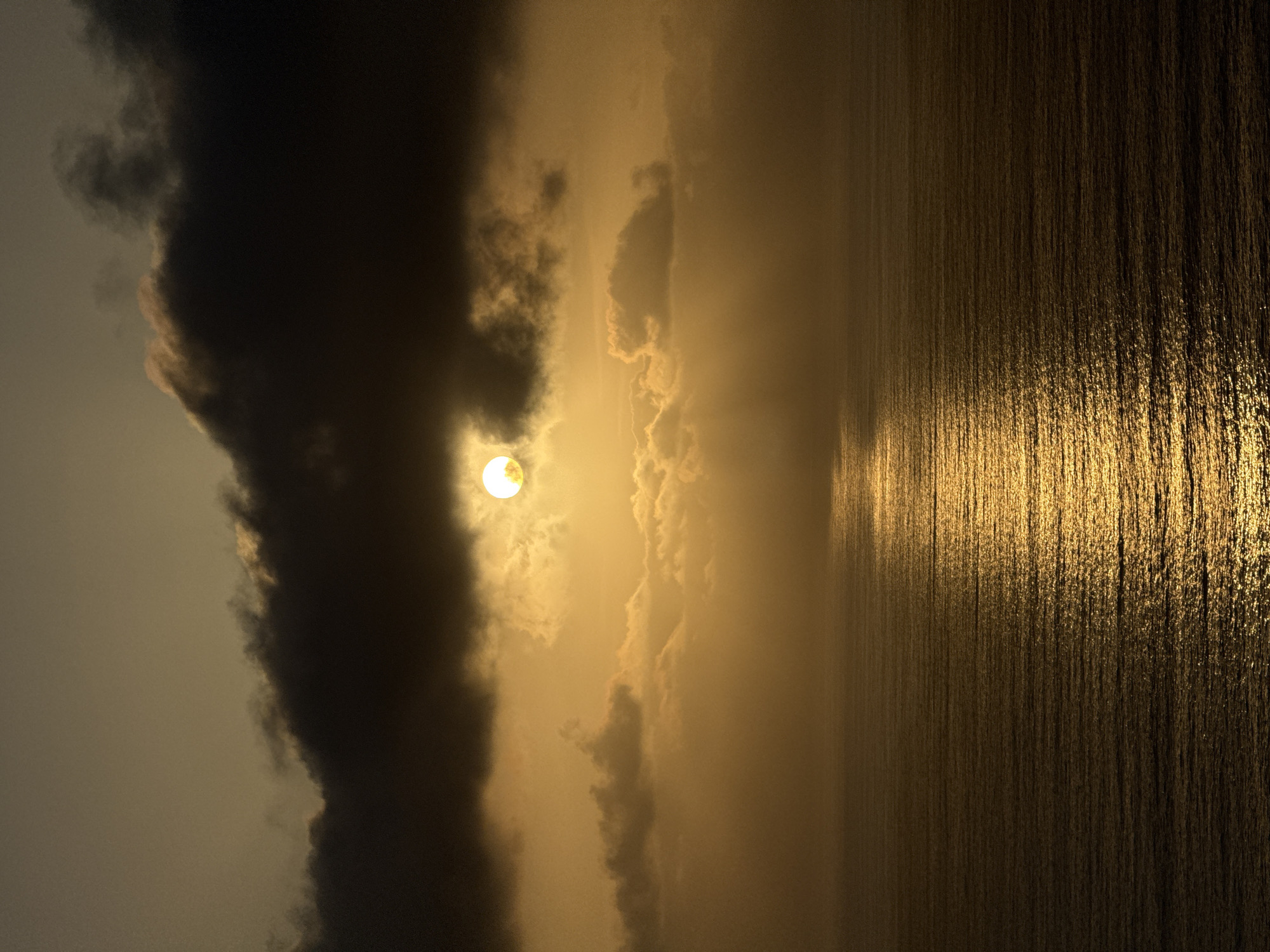 A scenic photograph captures a sunset over the Pacific Ocean in San Diego, California. The sun, partially obscured by dark clouds, casts a warm golden glow across the sky. Rays of sunlight break through the clouds, illuminating scattered lighter clouds below and creating a shimmering reflection on the water's surface. The ocean is calm, with gentle waves rippling across its expanse. The foreground is dominated by the dark, textured surface of the water, while the background features the dramatic interplay of sunlight and clouds. The image is rich in warm tones, ranging from deep browns and grays in the clouds to bright yellows and golds in the sunlight and its reflection.