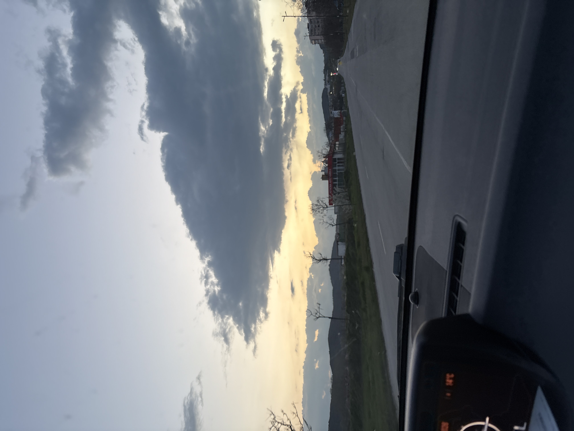 The image depicts a view from inside a vehicle driving along a road, presumably in Panagyurishte, Bulgaria. The road has visible lane markings and is flanked by greenery and trees on one side, with buildings in the distance. Above, the sky is partially obscured by dramatic, heavy clouds with a visible sunset illuminating the lower parts. A portion of the vehicle's dashboard is in the lower right corner of the frame, displaying temperature of 24°C.