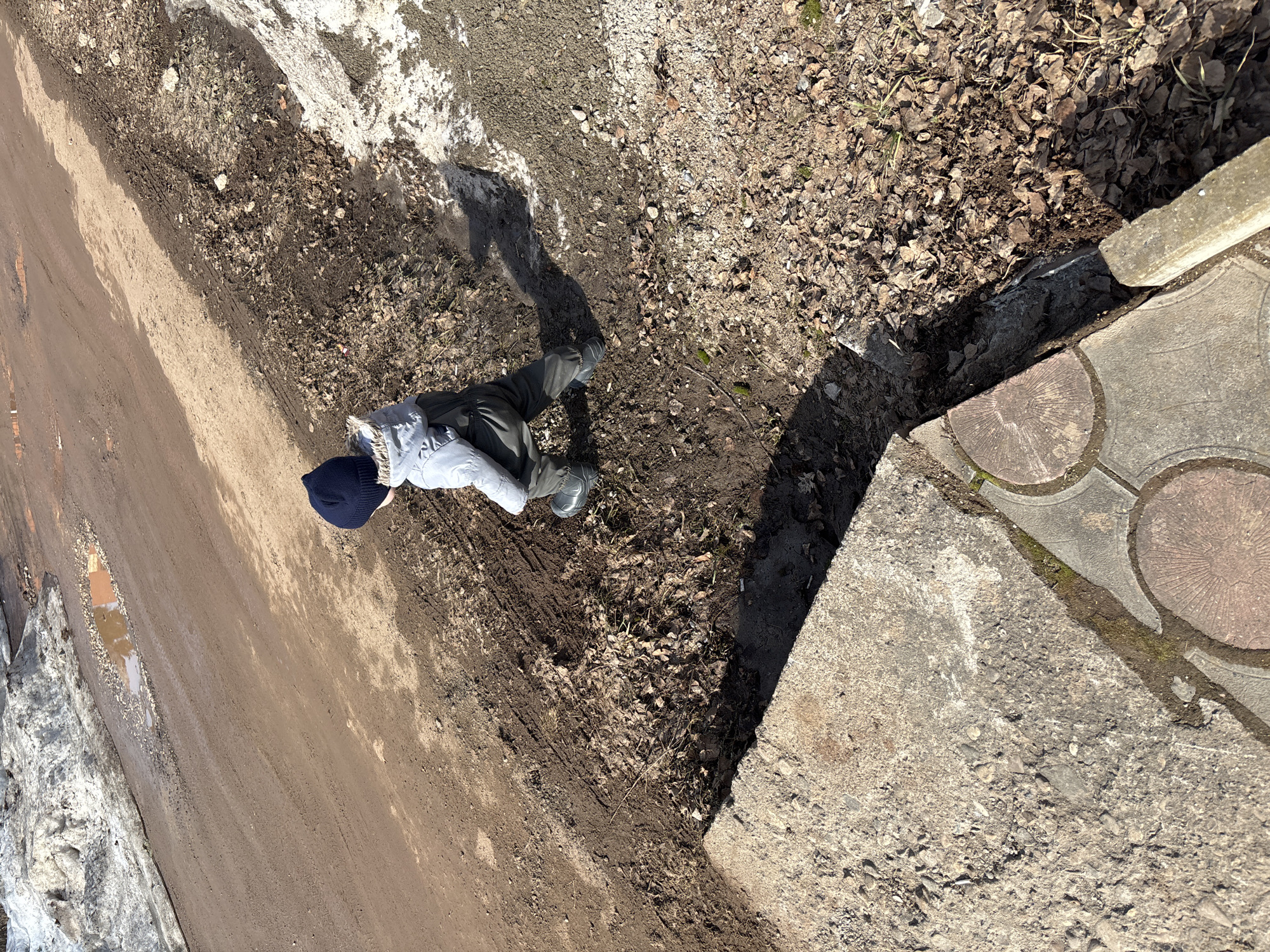 A high-angle shot depicts a person standing on a dirt patch adjacent to a road in Ферма, Russia. The person is wearing a dark blue beanie, a light-colored jacket, dark pants, and dark-colored boots. They are casting a shadow. The ground consists of mud, dirt, and dead leaves. To the right, there's a concrete structure with patterned pavement stones. The road is muddy and bordered by melting snow and ice. A puddle is visible on the road.