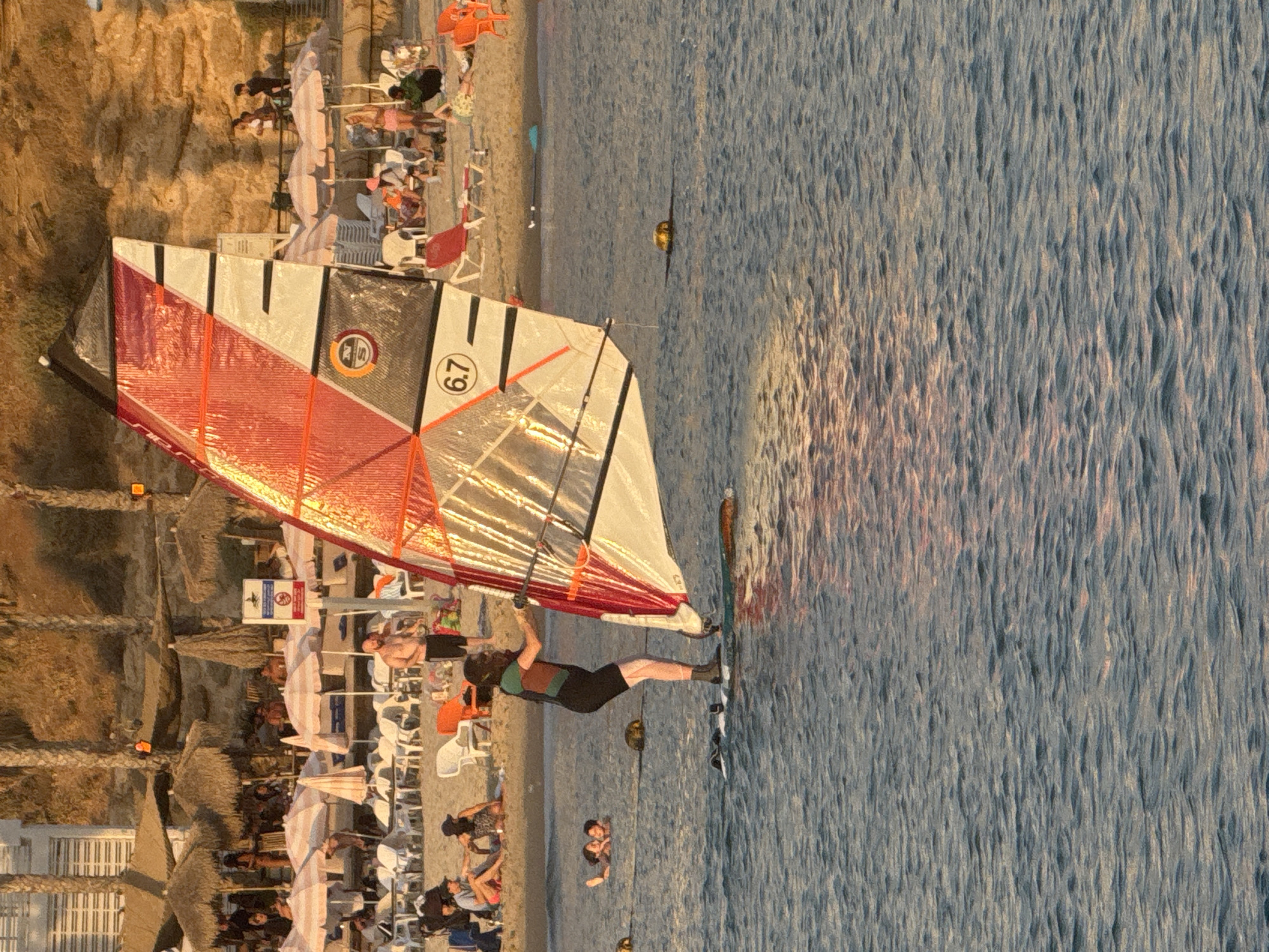 The image depicts a windsurfer in the foreground on the water, with a busy beach and cliff face in the background under golden hour lighting. The scene is located on a beach in Tel Aviv, Israel.
In the immediate foreground, a person is actively windsurfing. The individual wears a dark wetsuit or rashguard with a teal/green section on the torso, dark shorts, and dark water boots. They are standing on a dark-colored windsurf board and gripping the boom of a large sail. The sail is predominantly red, white, and black, featuring reflective material. A circular logo with an "N" and "S" appears on the sail, along with the number "6.7". To the left of the windsurfer, two individuals are partially submerged in the water, appearing to be swimming or floating. Several yellow buoys are spaced across the water surface. The water exhibits small ripples, reflecting warm, reddish-orange light on the right side of the frame, indicating the position of the sun.
The midground features a sandy beach densely populated with numerous individuals. Many people are seated on white plastic lounge chairs, with some red lounge chairs also visible. Various beach umbrellas are present, including multiple thatched straw umbrellas and striped fabric umbrellas, primarily pink and white. A large blue sign with white symbols and a red circular warning sign are positioned near the center of the beach area. People are engaged in various leisure activities, including sitting, walking, and conversing.
The background consists of a steep, sandy, and rocky cliff face. Sparse green vegetation, including several palm trees, grows along the top and face of the cliff. Structures resembling multi-story buildings with horizontal window patterns are visible atop the cliff, suggesting urban development. Additional people can be seen standing on a promenade or elevated area at the top of the cliff, observing the beach below. The overall lighting is warm, indicative of late afternoon or early evening, casting soft illumination on the scene.