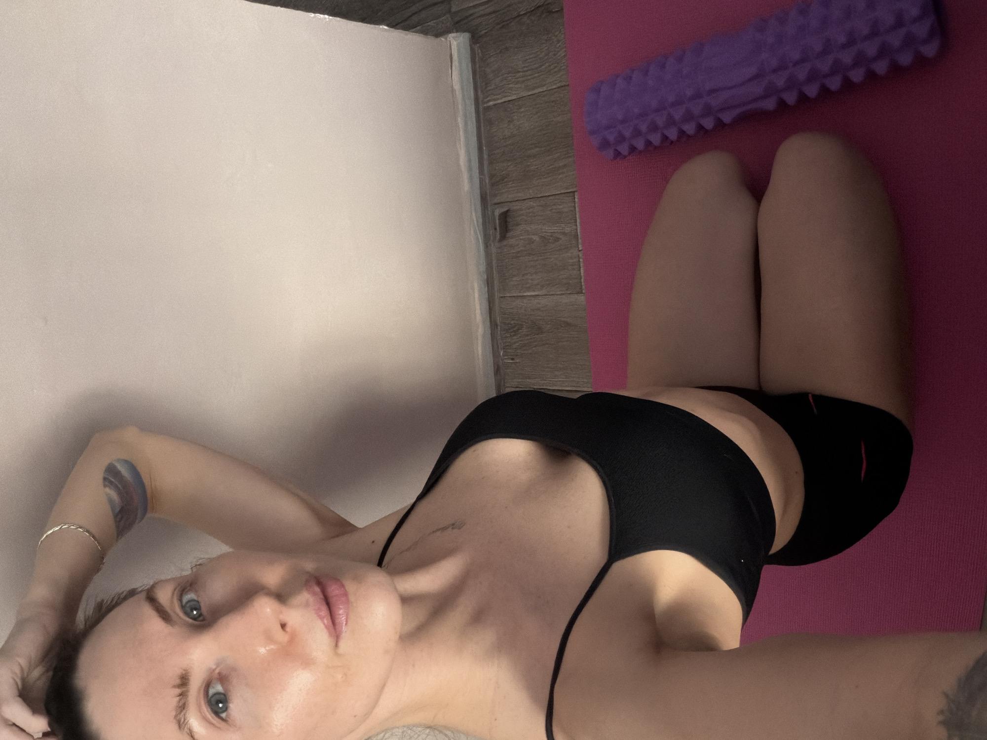 A woman with fair skin, blue eyes, and pink lips lies on a pink yoga mat with her head towards the bottom left corner of the image. She is wearing a black sports bra and black shorts. Her right arm is above her head and visible in the top left corner, bearing a circular, multi-colored tattoo and a silver bracelet. A purple foam roller rests at the top of the yoga mat, parallel to her knees. The background includes a light-colored wall and a section of light wood flooring. The location appears to be indoors. The city of Perm, Russia is not mentioned or implied in the image.