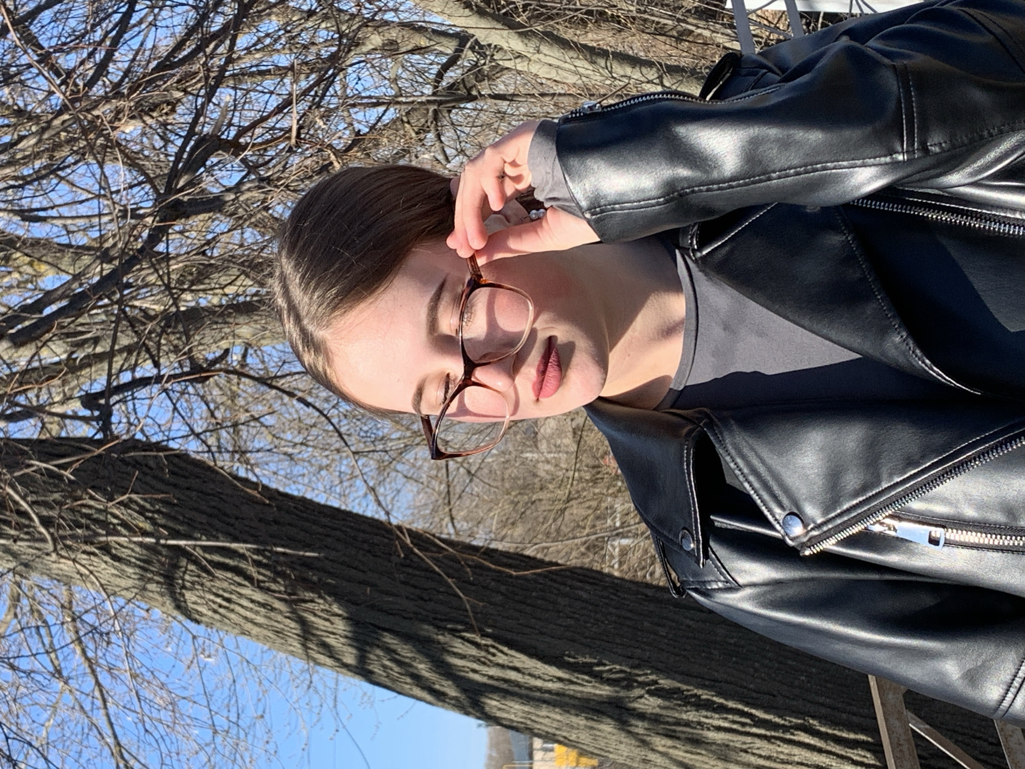 A medium shot captures a young woman outdoors on a clear, sunny day, likely in a natural setting such as a park in Фоки, Russia. The woman, positioned slightly off-center to the right, is seen from the chest up. She has medium-brown hair and wears round eyeglasses with brown/tortoiseshell frames. Her expression is serene, with her eyes partially closed and lips slightly parted, displaying pink lipstick. She is dressed in a black faux leather jacket, featuring prominent zippers and snap buttons, worn over a grey long-sleeved shirt. Her right hand is gently raised, with fingers lightly touching the frame of her eyeglasses.
The background consists of a clear, bright blue sky and a dense network of bare tree branches, indicating a season without foliage such as late autumn, winter, or early spring. A large tree trunk with textured bark is visible in the lower-left and central parts of the frame. The scene is brightly illuminated by natural sunlight, casting distinct highlights on her jacket and face. No discernible text is visible in the image.