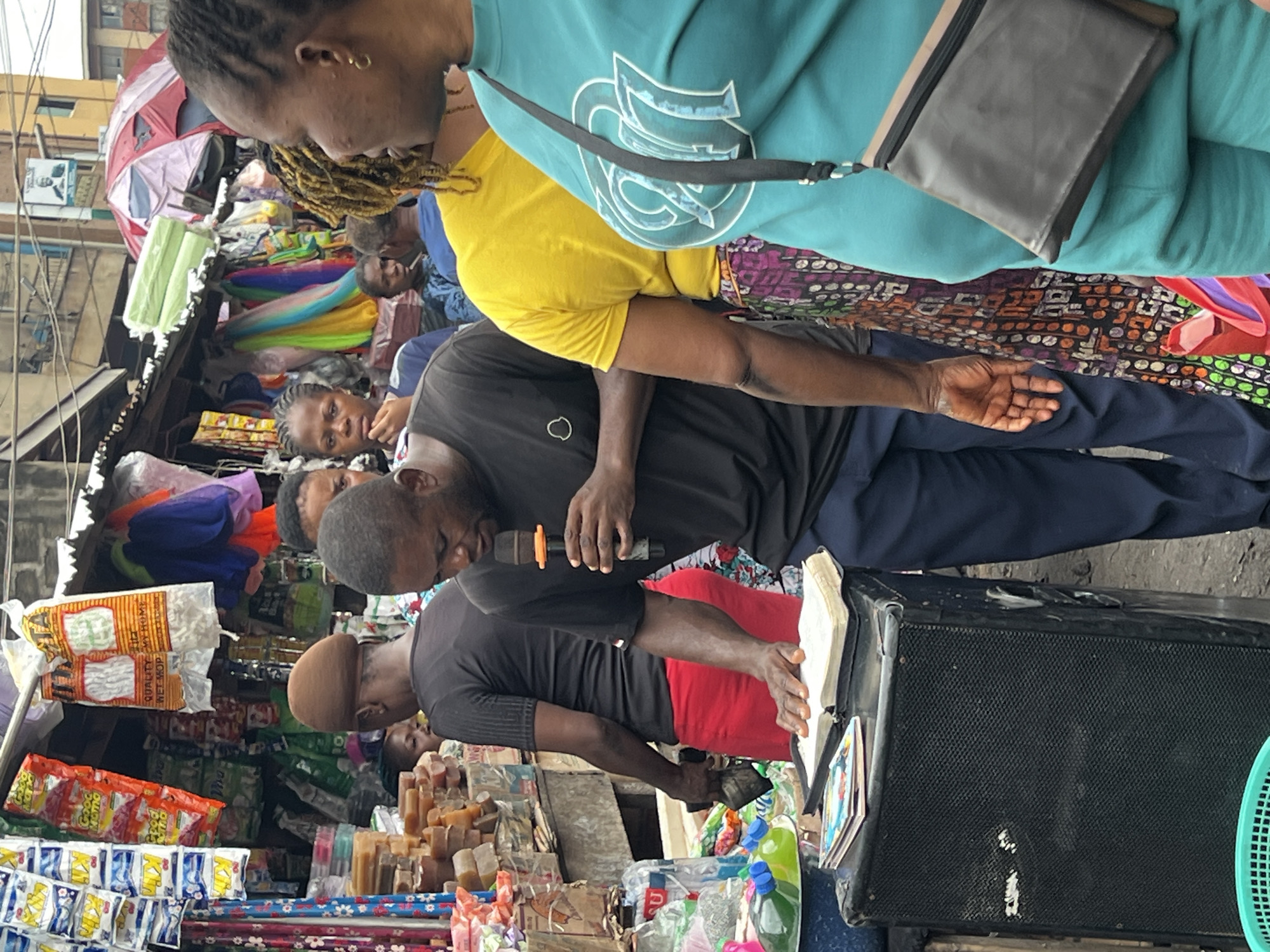 This image depicts a busy marketplace scene in Nkpor, Nigeria, likely during daylight hours given the general illumination. The foreground features several people, with a man in a black t-shirt and red shorts holding a microphone and speaking, possibly to a gathering. He is positioned near a large black speaker and a book, suggesting a sermon or public address.
Behind him, other individuals are visible, including a woman in a teal top and a man in a yellow shirt, both appearing to be engaged in market activities. The background is filled with stalls laden with various goods. Colorful textiles, fabrics, and packaged items are prominently displayed, indicating a vibrant commercial environment. Some of the packaged items have visible text, including what appears to be a brand name like "Good Mama" on some red bags, and other illegible text on white packaging and various colorful bags.
The overall atmosphere suggests a lively and bustling community market, with a focus on religious or public discourse by the man with the microphone. The image is oriented vertically, suggesting it was taken in portrait mode.