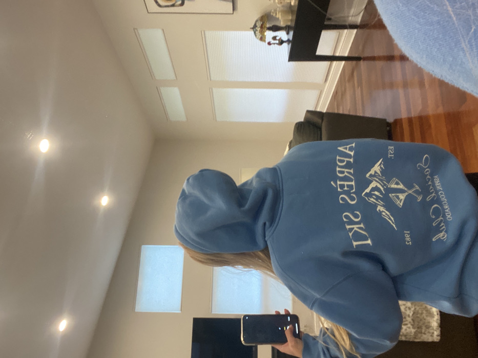 The image depicts an indoor residential scene in Ridgefield, United States, captured via a mirror reflection. The primary subject is a person, viewed from behind, wearing a light blue hooded sweatshirt. The back of the sweatshirt features an inverted white graphic design, which includes the mirrored text "APRÈS SKI" above a mountain range, crossed ski poles or cocktails, and the mirrored text "Social Club EST. 1985 ASPEN COLORADO." The subject holds a dark-colored smartphone in their left hand, positioned as if taking the photograph.
The environment includes light beige walls and a vaulted ceiling with three visible recessed lights. The flooring is dark hardwood. On the left, a large, dark flat-screen television is visible. In the background, a dark gray sofa or sectional is present. On the right, a dark wooden console table holds a Tiffany-style stained-glass lamp, a red rooster figurine, and several white decorative objects. Multiple windows are present: two upper rectangular transom windows and two lower windows with horizontal white blinds are visible on the right wall. Similar windows are partially visible on the left wall.