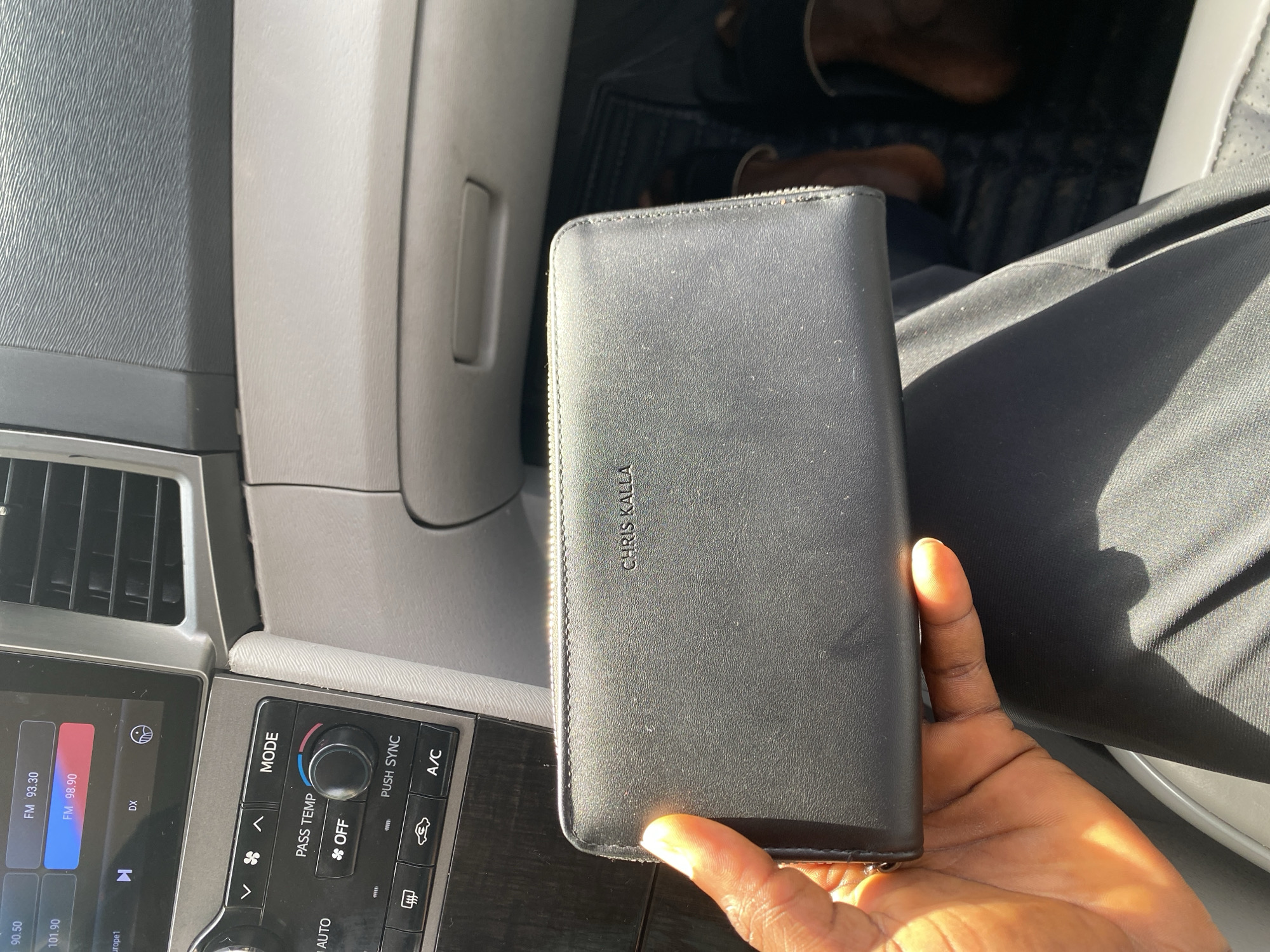 In Bar Jirgi Summa, Nigeria, a black leather wallet with the embossed name "CHRIS KALLA" is held by a person's hand. The wallet is positioned in the foreground, partially obscuring a car's dashboard which features various buttons and a screen displaying radio information. The interior of the car is visible, with parts of seats and flooring in the background. The lighting suggests a sunny day, with strong highlights on the wallet and hand.