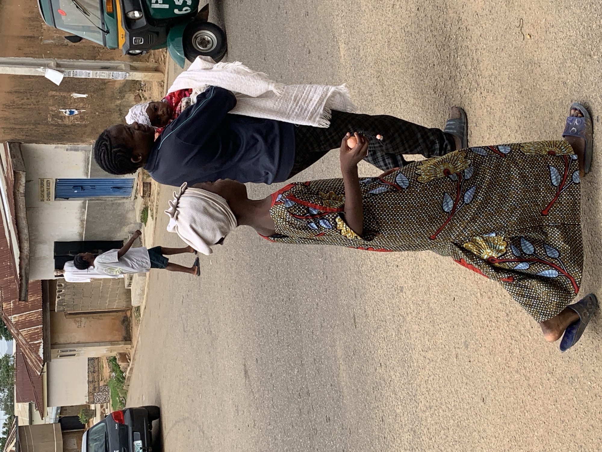 A daytime street scene in Jos, Nigeria shows a woman in a patterned brown and yellow dress and a white head covering holding an orange object. She is standing on the asphalt road, looking up at a man who is carrying a person wrapped in a white cloth. The man is wearing a dark blue top and dark trousers. A child with a white head covering is visible peeking out of the cloth. A young man walks away from the group in the background. A green vehicle is partially visible in the upper left, and a black car is parked on the left side of the road. Buildings line the street, with a mix of brick and metal roofing visible.