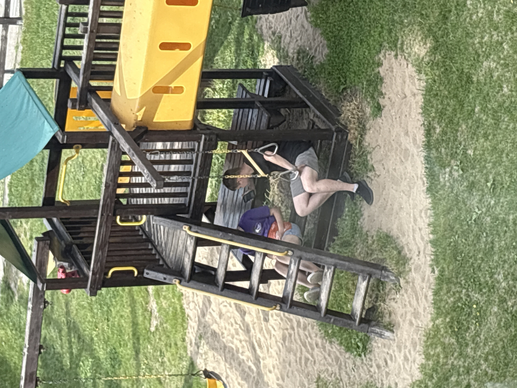 The image depicts a playground structure with two individuals. One individual is hanging from a rope swing within the structure, with their feet dangling. Another individual sits below on a set of wooden steps of a ladder, their body positioned beneath the first individual. The playground is surrounded by grass and a sandy surface. The location is outdoor. The structure has a yellow slide and a small green roof.