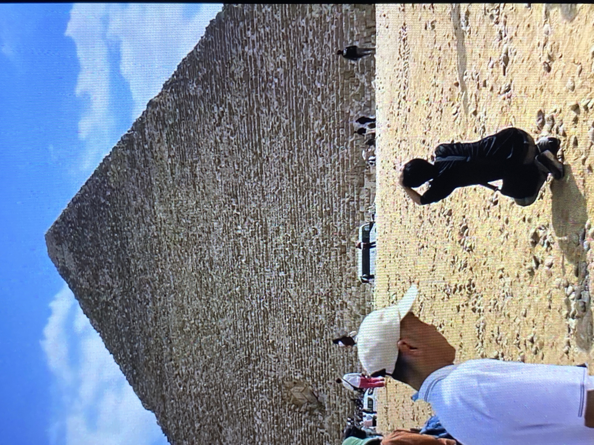 A pyramid stands prominently against a blue sky with clouds. In the foreground, several people are visible. One person in a white shirt and a white baseball cap looks up at the pyramid. Another individual kneels on the sandy ground, their head bowed, possibly photographing or examining something. The surrounding terrain appears to be sandy and uneven. Other people and what appear to be tour buses or vehicles are also nearby. There is no evidence to support this image being taken in Tinaquillo, Venezuela. The pyramids are typically located in Egypt.