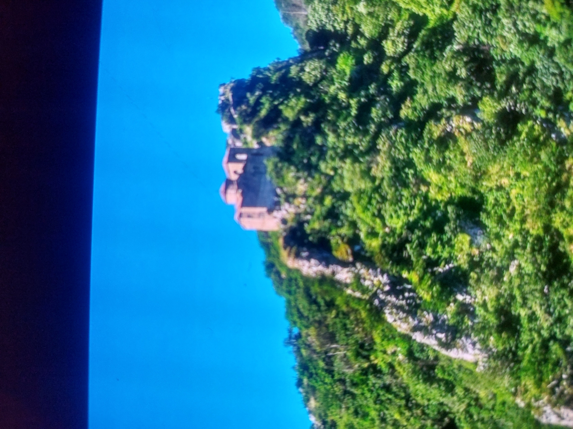 Here is a description of the image:
The image shows a rocky cliffside covered in lush green vegetation. At the top of the cliff, there appears to be a stone building with a red dome or roof. The sky is bright blue and fills most of the left side of the frame. There is a dark edge visible on the left, possibly indicating the edge of a screen displaying the image. The scene is possibly of a landmark in Tsarevo, Bulgaria.