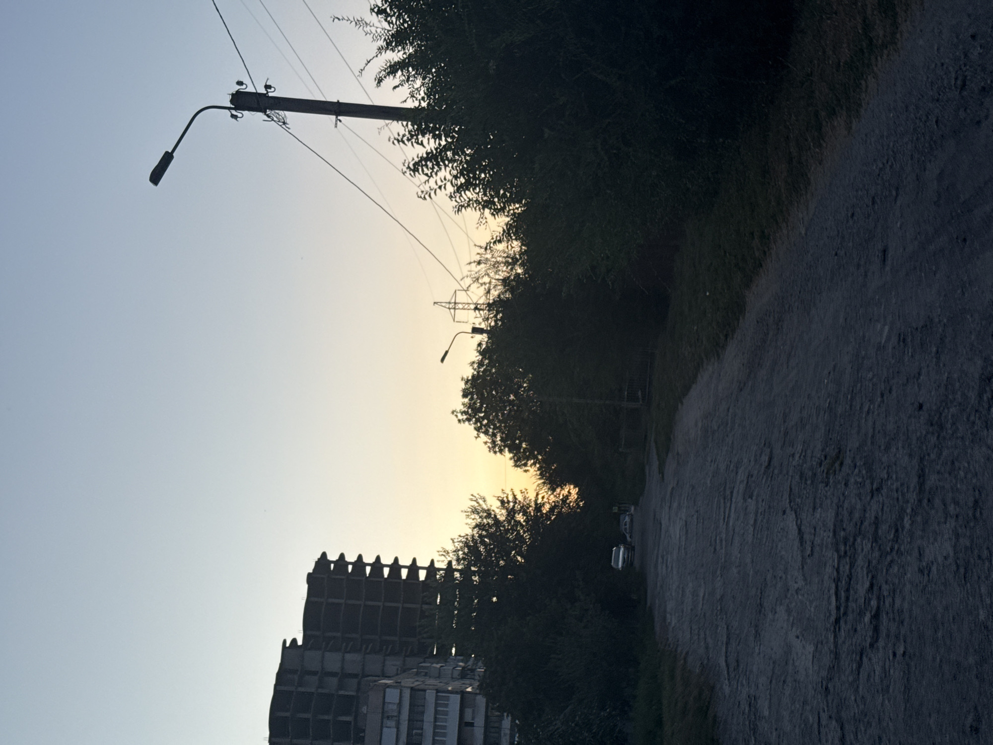 The image depicts an unpaved road in a city, possibly during dusk. To the left, an unfinished, multi-story concrete building is visible, with exposed structural elements and windows. Adjacent to it are trees and some grass. The road surface is uneven and appears to be made of gravel and dirt.
Further down the road, two cars are parked or stopped. On the right side of the road, a mature tree line forms a dense barrier. In the background, a streetlamp on a pole is illuminated, and power lines are strung across the sky. The sky exhibits a gradient of colors, from a pale blue at the top to a yellowish-orange hue near the horizon, indicative of sunset or sunrise. The overall impression is one of an urban fringe or an undeveloped area within a city. The presence of the unfinished building and the unpaved road suggests ongoing construction or a neglected section of the city.