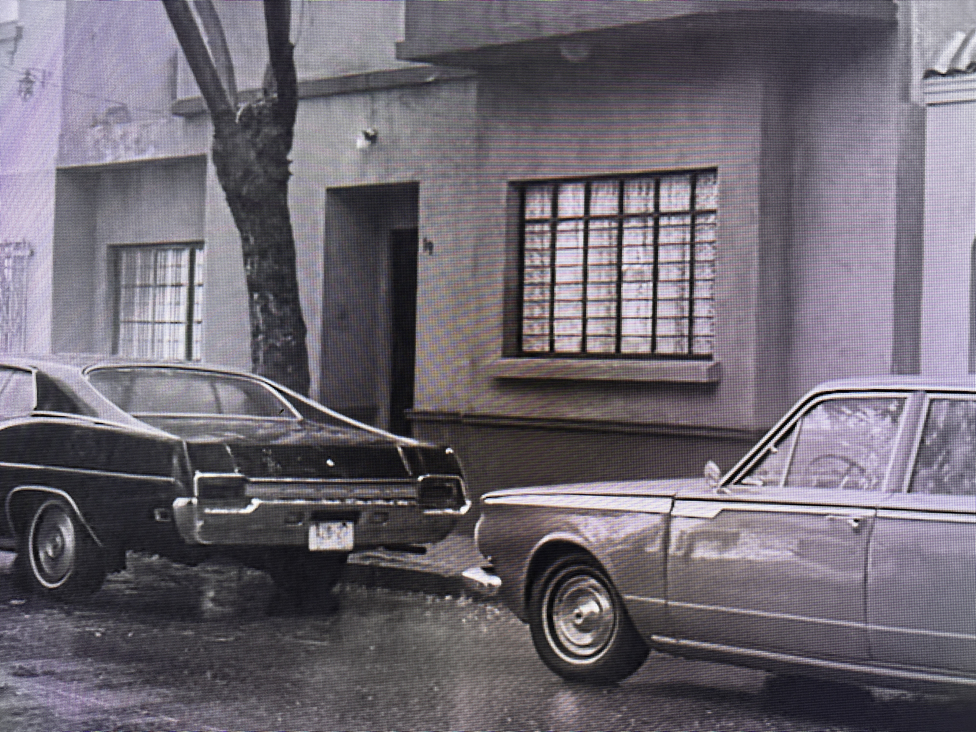 Two vintage cars are parked on a wet street in front of a building in Alaverdi, Armenia. The car on the left is a dark sedan, and the car on the right is a lighter-colored sedan. The building appears to be a multi-story structure with windows and a doorway. A tree is visible on the left side of the image. The scene is captured in black and white.