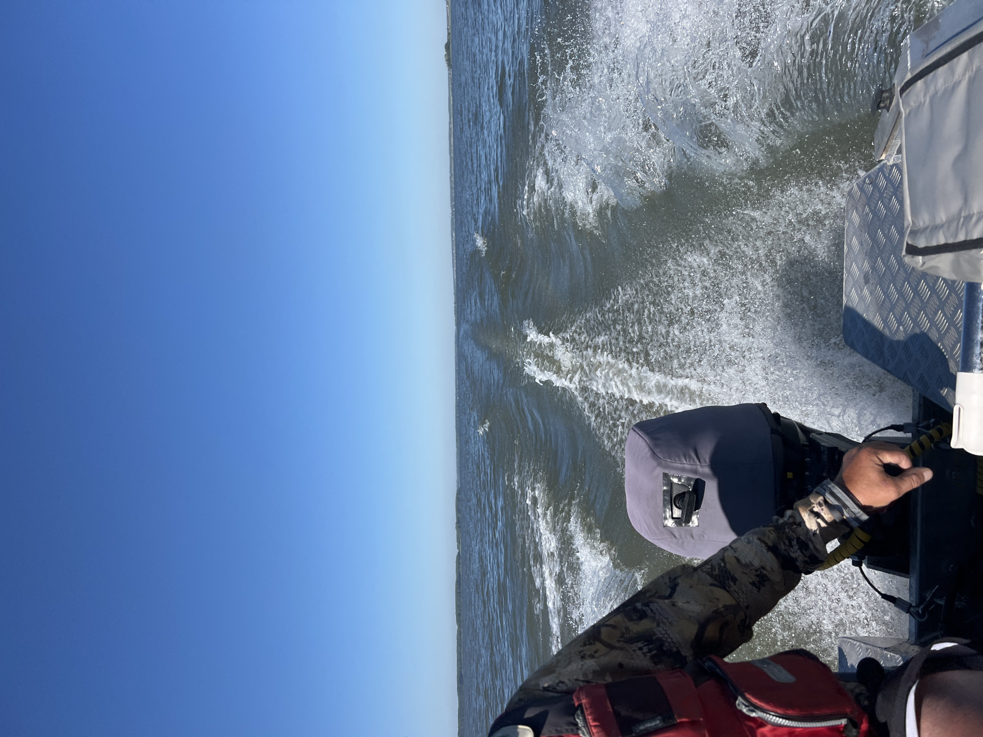 A man wearing a camouflage long-sleeved shirt, a red life vest, and black shorts, is steering a boat on a body of water under a clear blue sky. He is holding the steering mechanism of the boat's outboard motor with his right hand. The motor is covered with a gray canvas. Water is churning and splashing behind the boat, creating a wake. In the distance, a thin line of trees is visible on the horizon, suggesting land. The image appears to be taken from the perspective of someone sitting behind the person steering. The location is not specified in the image or provided information beyond it being in the city of Beregovoe, Russia.