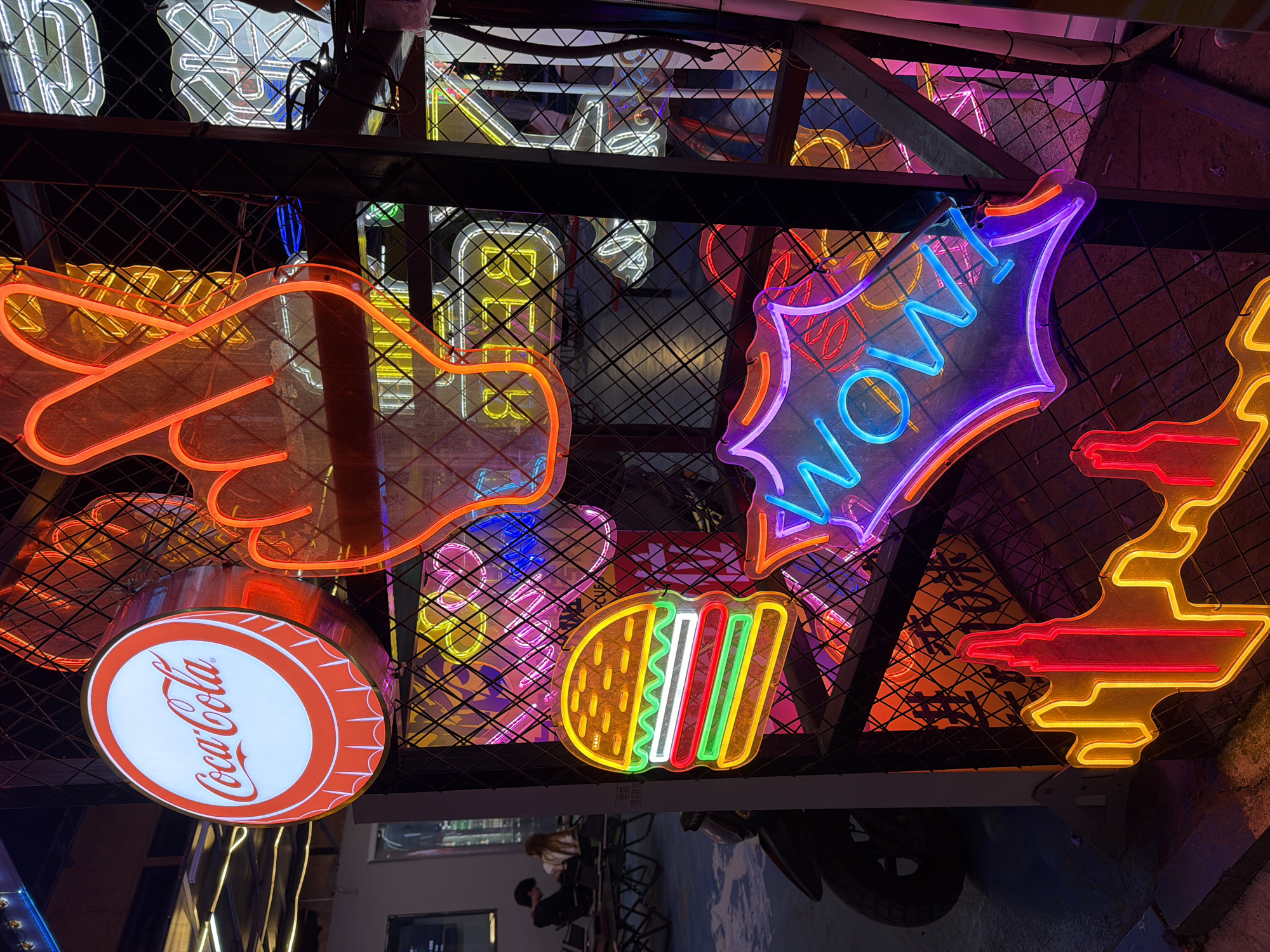A vibrant display of neon signs hangs in Chongqing High-tech Zone, China. The foreground features a Coca-Cola bottle cap sign, a pointing hand gesture, a "WOW!" speech bubble, a hamburger, and abstract shapes. The signs are mounted on a metal grid. In the background, blurred figures are visible through a window, along with parts of a vehicle and urban setting.