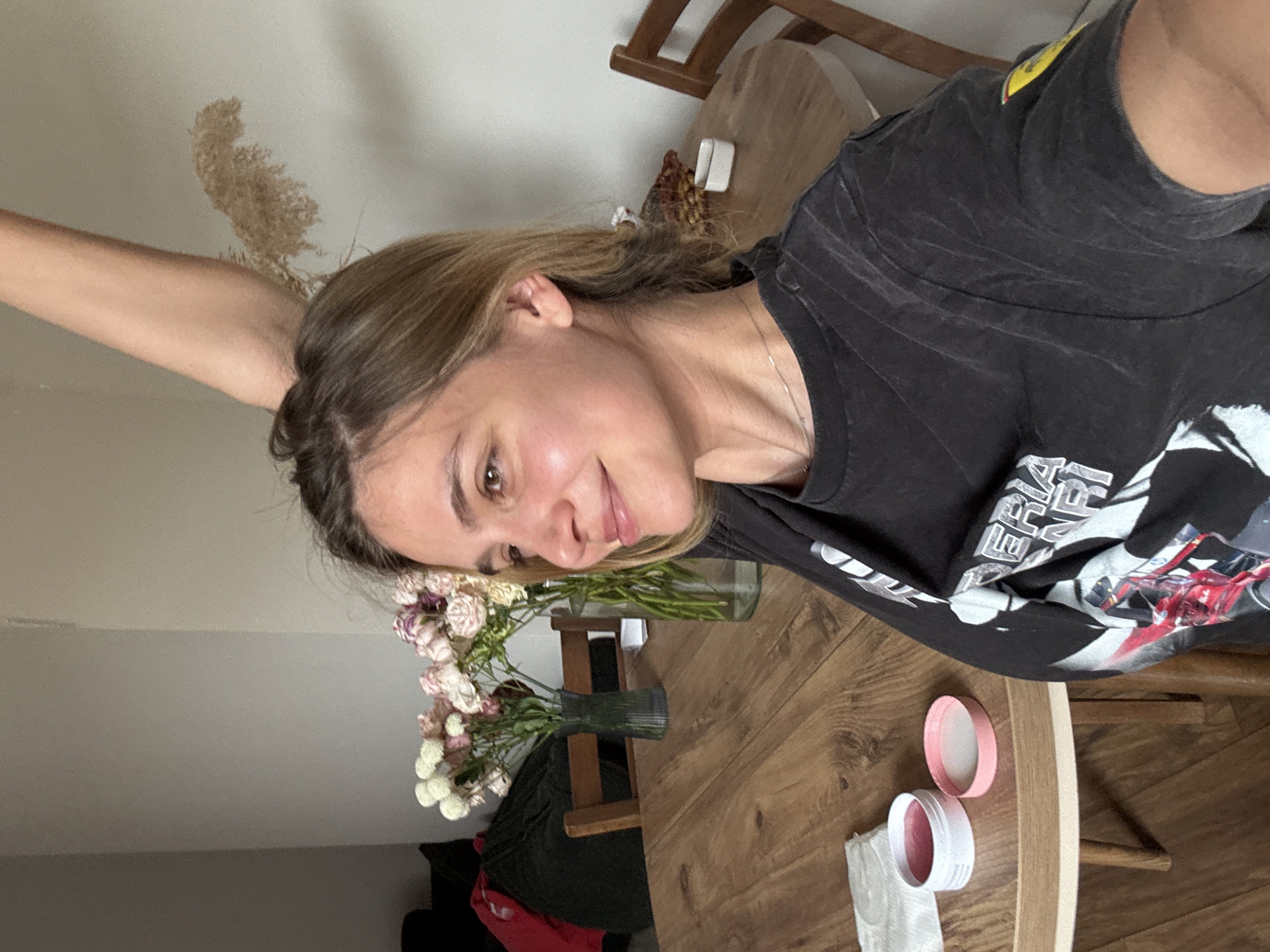 The image shows a fair-skinned person with light brown hair, wearing a dark-colored t-shirt. The person appears to be in a domestic setting, with a wooden table in the foreground displaying vases of flowers, one containing white and pink carnations and the other containing green foliage. On the table are also a white container with a pink lid, and a white tissue. A wooden chair and a portion of a black and red garment can be seen beneath the table. The setting seems to be indoors, against a light gray wall. The location is Unknown, Unknown.
