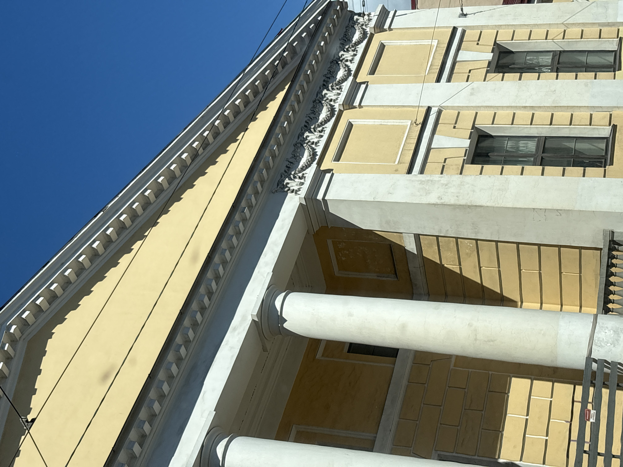 The image shows the facade of a pale yellow building with neoclassical architectural elements against a clear blue sky. The building features large white columns, a decorative cornice with what appears to be a garland motif, and rectangular window openings. The sunlight casts shadows across the building's textured surface. In the foreground, a dark railing is partially visible. The building's style and architecture suggest it is likely located in the historic center of Saint Petersburg, Russia.