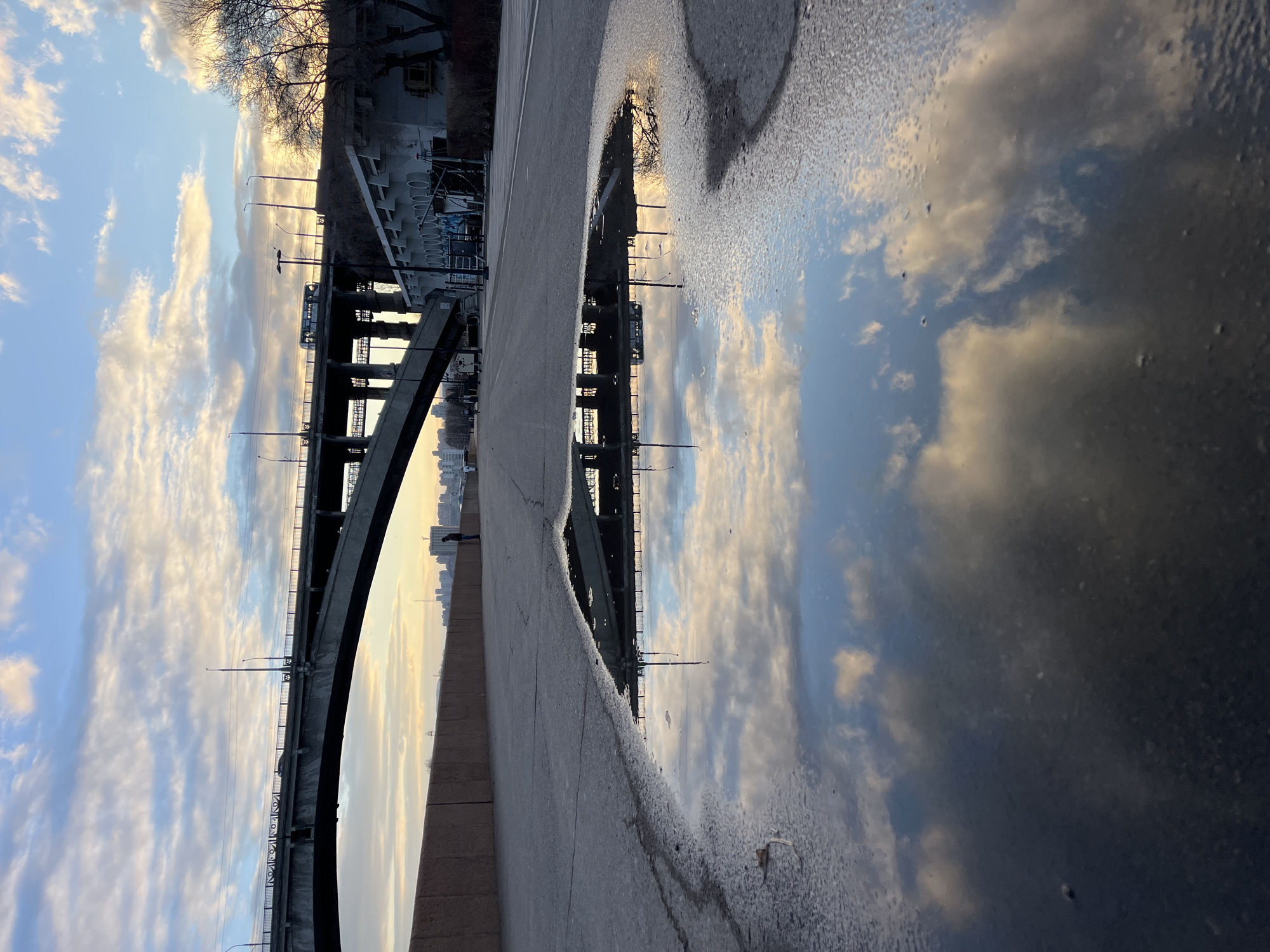 The image captures an urban landscape featuring a large bridge and a prominent reflective puddle. The scene is viewed from a paved area, looking towards a body of water.
In the foreground, a substantial, irregular-shaped puddle occupies the right side of the frame, reflecting the cloudy sky and parts of the bridge structure with remarkable clarity. The surface of the puddle shows slight texture, potentially from small ripples or ice formation.
A robust bridge, likely constructed from concrete or steel, extends horizontally across the middle of the frame. It features a distinct arching support structure, with multiple vertical and diagonal supports visible beneath the bridge deck. Streetlights are mounted along the bridge's edge, and power lines or cables run above it.
The sky, visible above and reflected in the puddle, is adorned with scattered, soft clouds ranging in color from white to golden, set against a pale blue backdrop. This warm, golden light near the horizon suggests either a sunrise or a sunset. In the far distance, beneath the bridge, a faint, silhouetted city skyline is discernible. To the left, a bare-branched tree stands silhouetted against the sky. A lone figure of a person is faintly visible walking on the far side of the bridge in the distance.
The time of day is evidently either early morning or late afternoon, characterized by the soft, warm ambient light. The presence of the puddle indicates recent wet conditions. No visible text is present in the image. The setting conveys a serene atmosphere, common to a riverside urban environment like Krasnoyarsk, Russia.