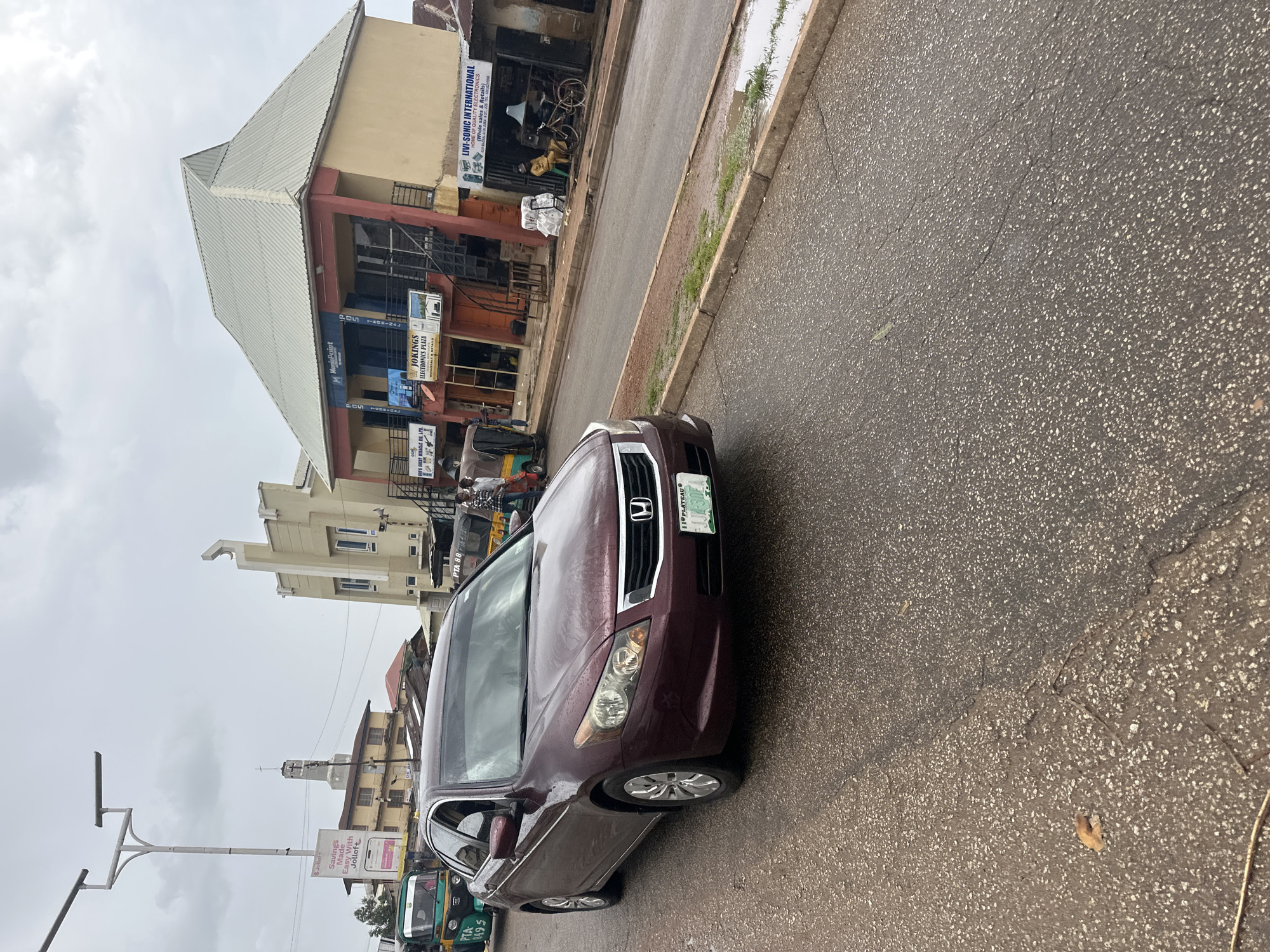 This media file captures a street scene in Jos, Nigeria, under overcast skies, suggesting it might be morning or afternoon. The wet asphalt of the road indicates recent or ongoing rain, contributing to a slightly dull atmosphere.
The primary focus is a maroon sedan parked on the side of the road, facing away from the viewer. Its headlights and front grille are visible. Beside it, several tricycles, a common mode of transport in Nigeria, are also present. One tricycle, green and yellow, is particularly prominent. In the background, a multi-story building with shops on the ground floor lines the street. Signs on the building advertise various businesses, including "LIVI-SONIC INTERNATIONAL HOME OF QUALITY ELECTRONICS" and "JOKINGS ELECTRONICS PLAZA." Another sign for "MoniePoint" and a promotional poster for "Jollof Savings" are also visible. A utility pole with streetlights stands to the left of the frame. The overall scene depicts a typical urban commercial area in Nigeria, likely during a period of rain.