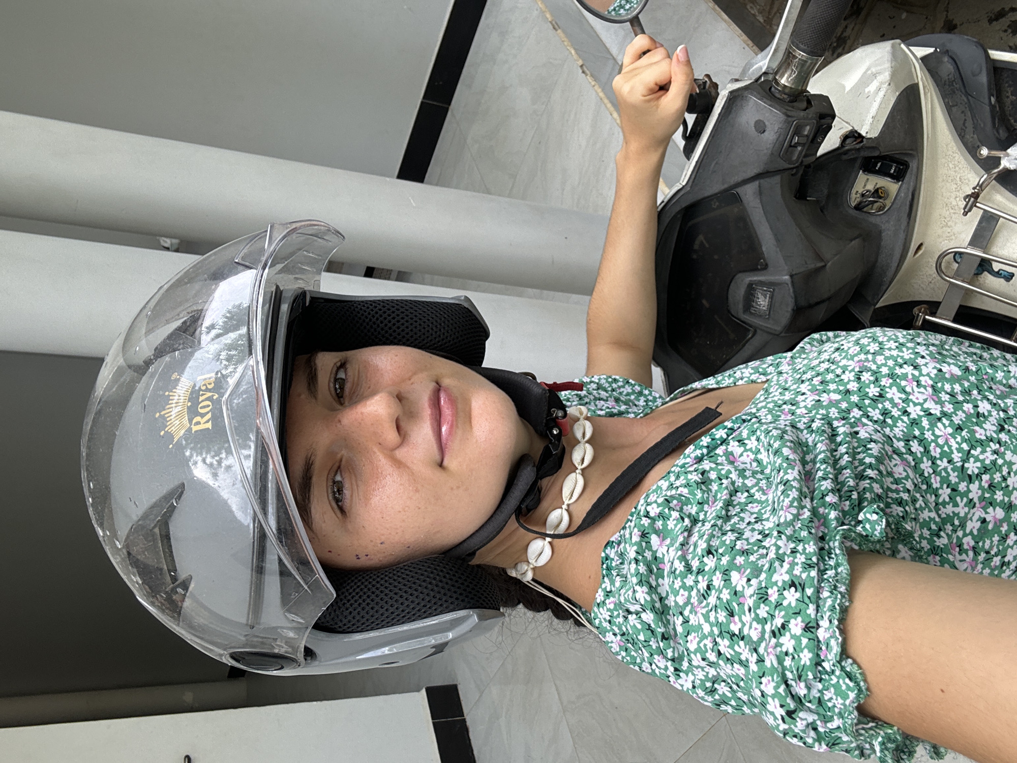 A young woman wearing a full-face motorcycle helmet and a green floral-patterned dress is seated on a scooter. She has a necklace made of seashells. Her right arm is extended, holding the scooter's rearview mirror. The scooter is parked in a paved area with a white pillar in the background. This image was likely taken in the city of Mũi Né, Vietnam.