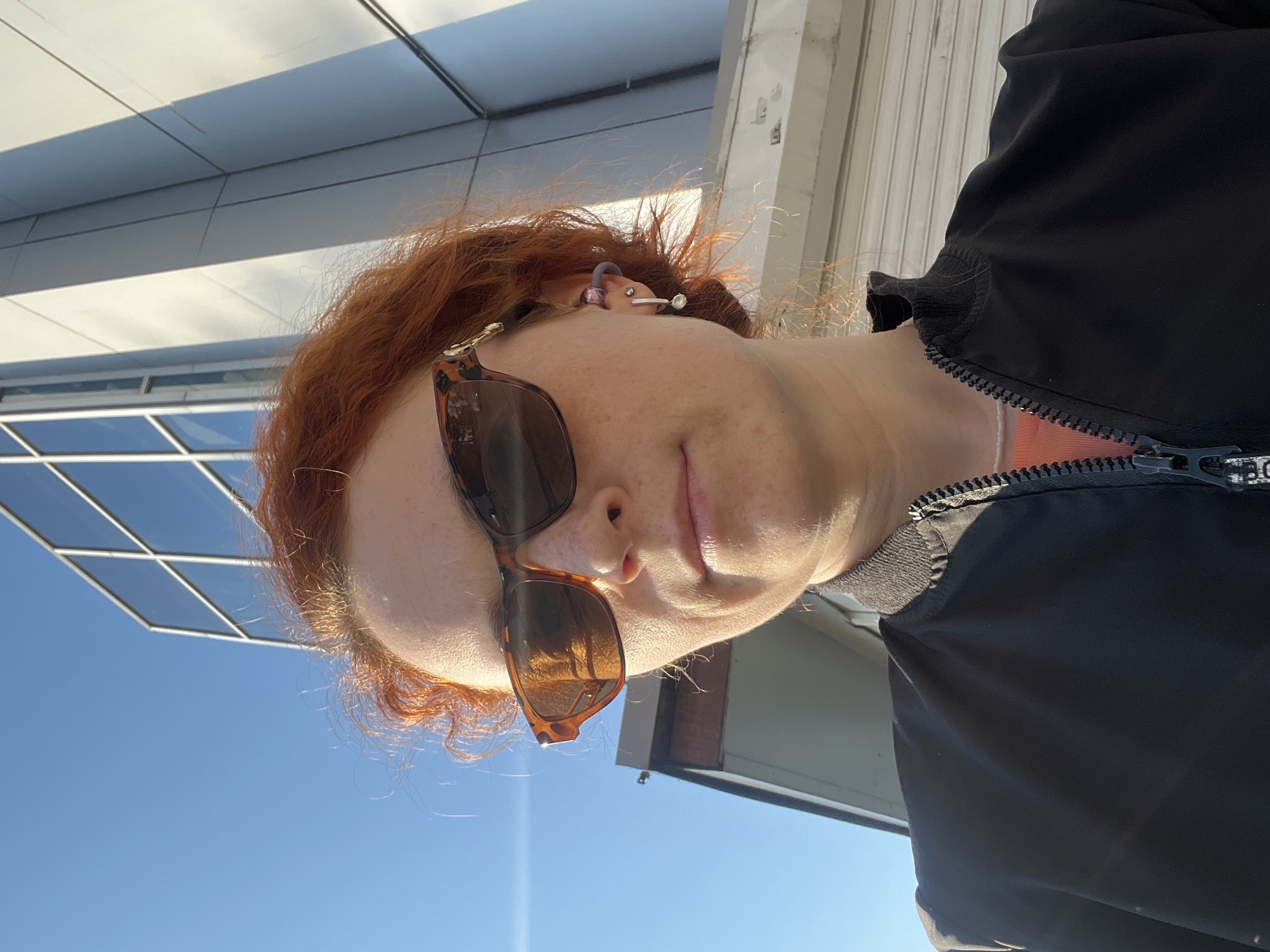 A woman with red hair is wearing sunglasses and a black jacket. She is outside on a sunny day, and a modern building with glass windows is visible in the background. The location is likely in an urban area, but the specific location in Chaikovsky, Russia cannot be determined with certainty.