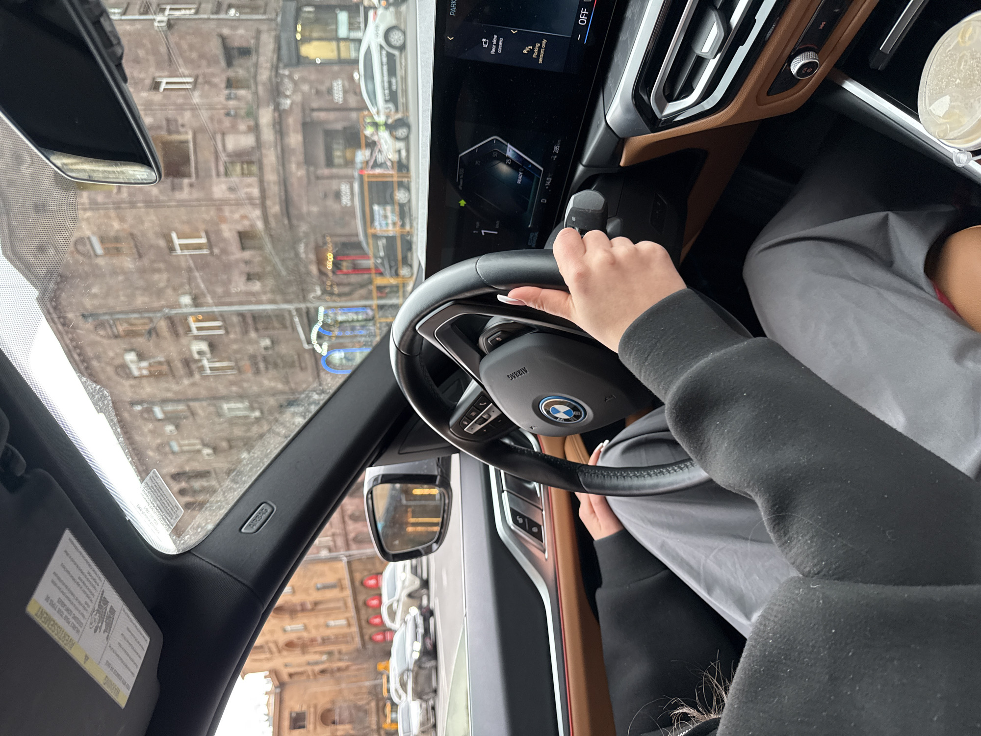 The interior of a car is captured, displaying a person’s hand gripping the steering wheel, marked with the BMW logo. The car features a digital dashboard with illuminated displays, as well as tan-colored accents along the dashboard. The driver wears a gray jacket. In the passenger area, a cup of liquid can be observed. Outside the windshield, a cityscape is visible, indicating an urban setting with densely packed buildings and parked vehicles. The urban environment suggests a location in the city of Yerevan, Armenia.