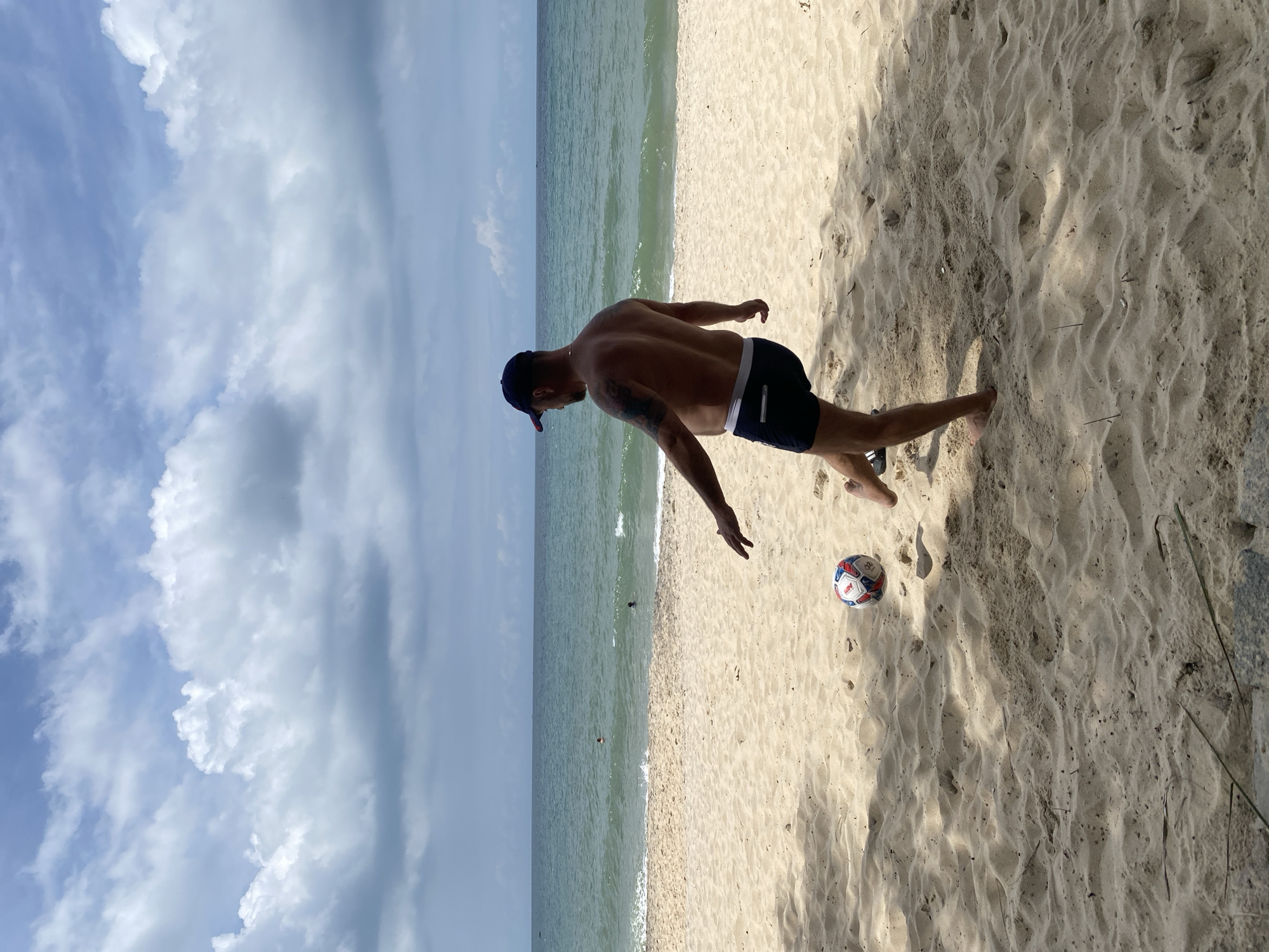 An adult male wearing dark swim trunks and a blue baseball cap is standing on a sandy beach, kicking a soccer ball. The man is facing away from the camera, his back to the viewer. He has tattoos on his left arm. The soccer ball is in mid-air, just in front of his right foot. The beach is covered in light brown sand, with some scattered debris. In the background, the turquoise ocean meets the horizon under a sky filled with dramatic white and gray clouds. There are a few people visible in the water in the distance, appearing to be swimming. The overall scene suggests a casual day at the beach.