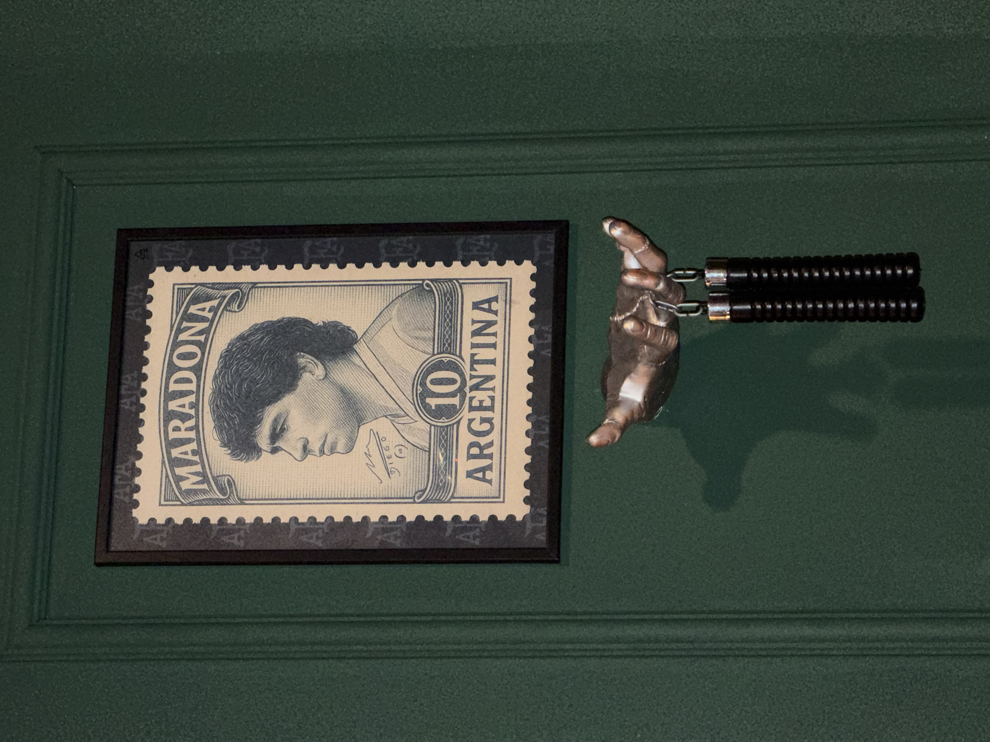 A framed stamp depicting Diego Maradona is mounted on a green wall with wainscoting. To the right of the frame, a metallic hand protrudes from the wall, holding a pair of nunchucks. The location is Sofia, Bulgaria.
