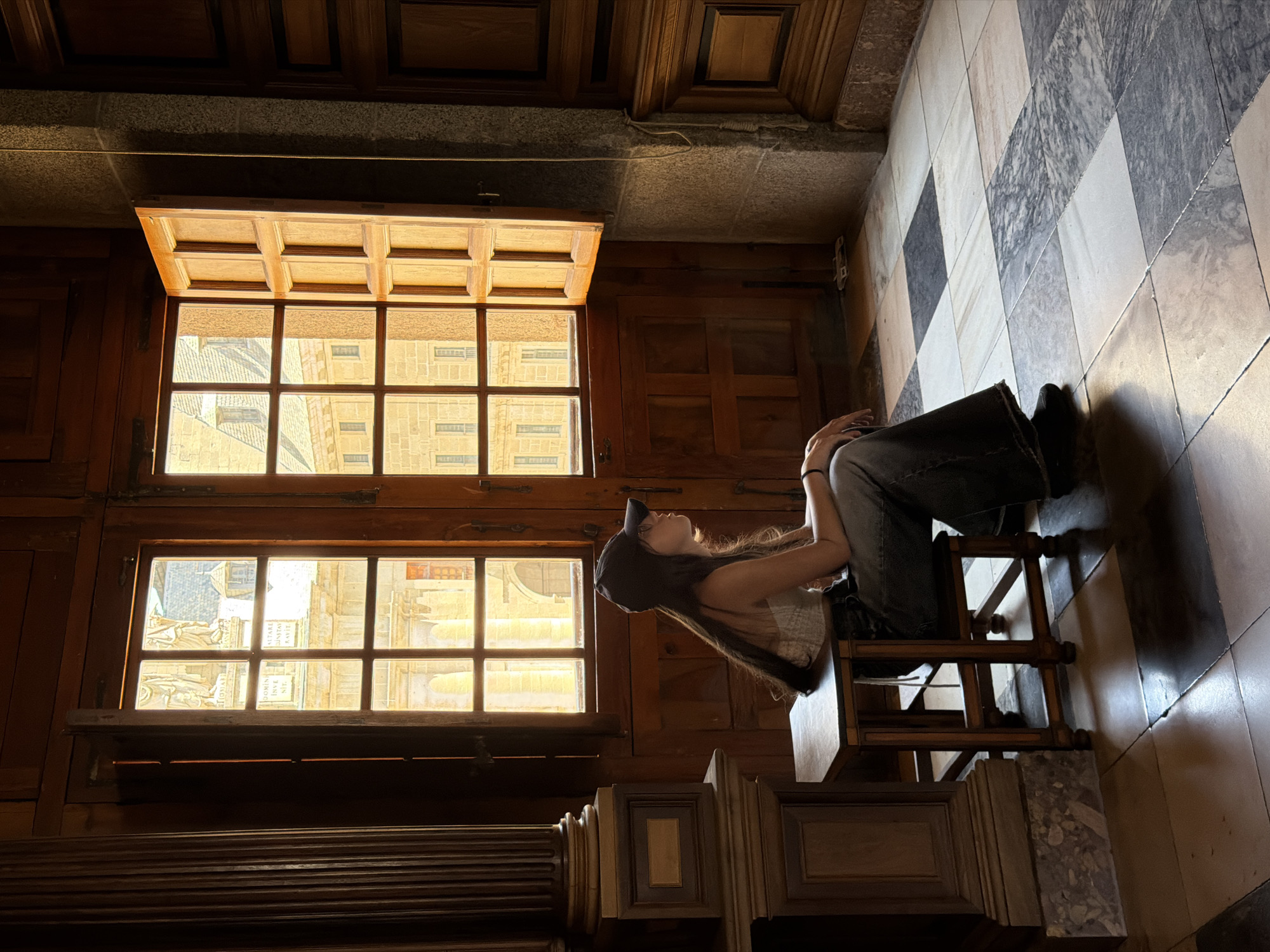 A young woman wearing a baseball cap, a light-colored tank top, and dark jeans sits on a wooden stool, looking upwards and to her right. She is positioned near two large, multi-paned windows that offer a view of an ornate building with a pale yellow facade. The windows themselves have wooden frames and grilles, and the lower window has a wooden shelf beneath it. The room features dark wooden paneling on the walls and ceiling. The floor is tiled in a diamond pattern of light and dark squares, with some marble-like veining in the lighter tiles. A large wooden architectural element, possibly a column or decorative piece, is visible in the lower left of the frame. The lighting suggests it is daytime, with sunlight streaming through the windows. The overall mood appears calm and contemplative.