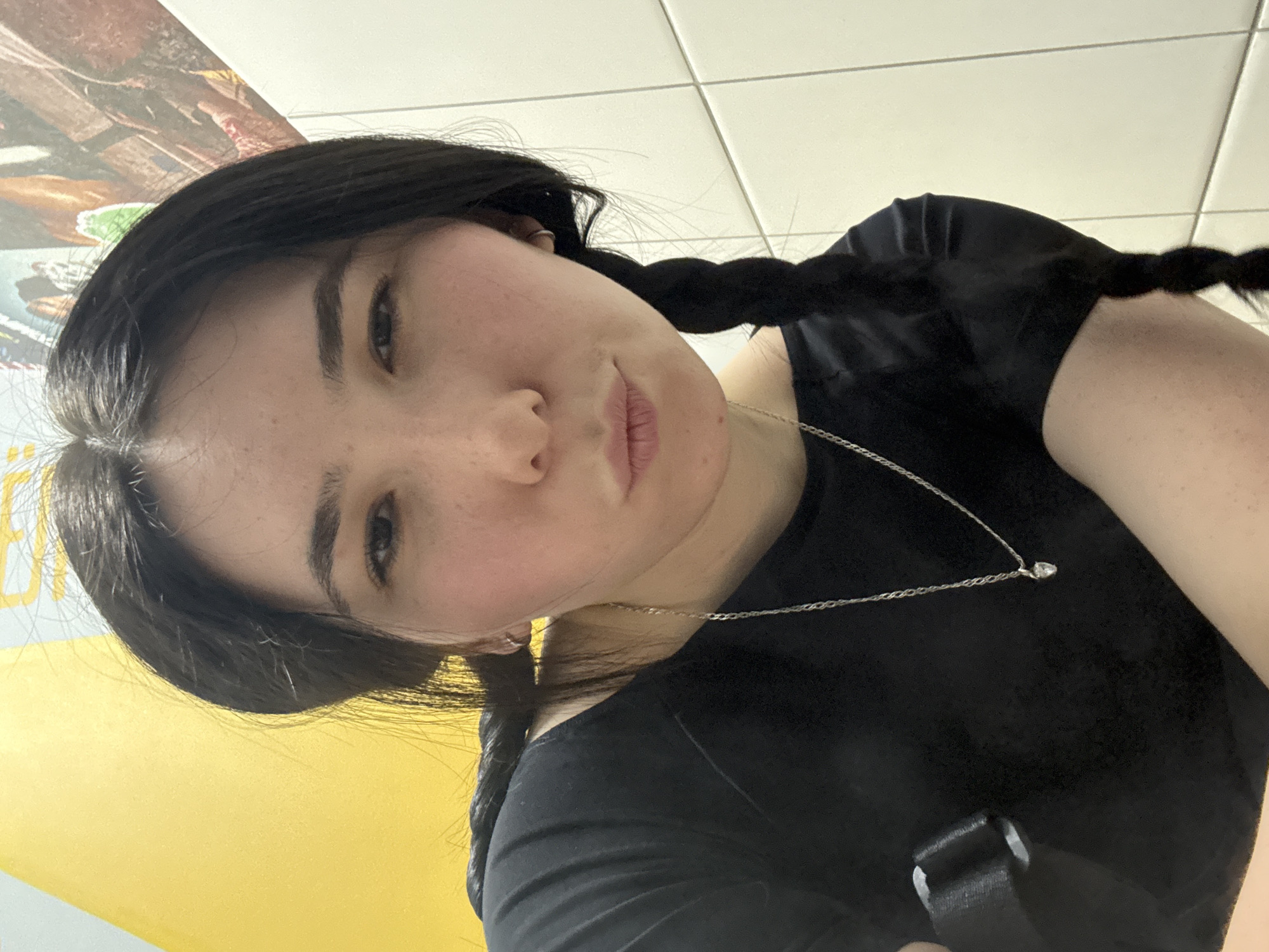 A young woman with dark hair in pigtails is posing for a selfie. She is wearing a black t-shirt and a silver necklace with a small pendant. The background appears to be a tiled wall and a yellow surface. The location is uncertain. There is no verifiable connection to Izhevsk, Russia based on the provided image.