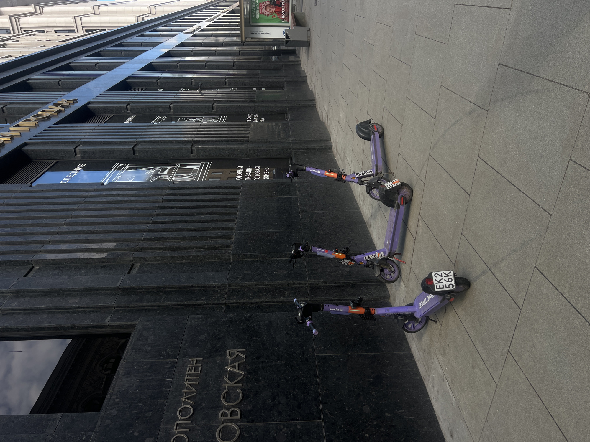 This photograph depicts a street scene in Moscow, Russia. In the foreground, three purple electric scooters with the "Юрент" logo are parked on a grey tiled sidewalk. The scooters bear license plates, including "EK2 56K" and "EE18 T". In the background, a building with a dark marble facade displays the inscription "ОПОЛИТЕН ОВСКАЯ" in Cyrillic, which translates to "Okhotny Ryad Metro Station". Window displays and commercial advertisements, some with Cyrillic text, are visible along the building's facade. A billboard advertising "ВКУСВИЛ" (VkusVill, a Russian supermarket chain) and a sign mentioning the "Мая" (Mayakovskaya) metro station can be seen above.