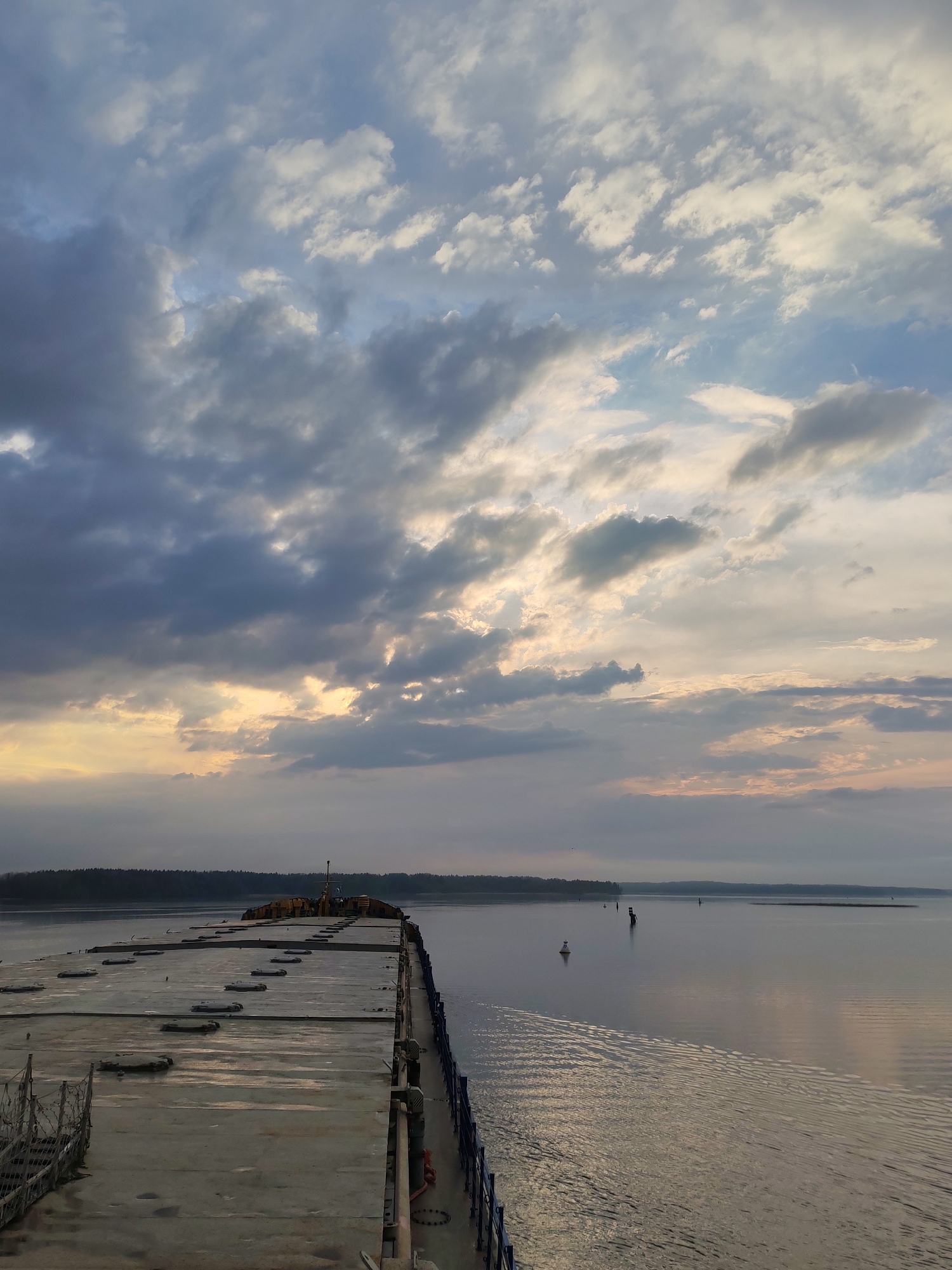 The image depicts a view from a large, flat-topped barge or ship on a body of water, likely a river or lake, with forested land visible in the distance. The sky is filled with clouds, some dark gray and some lighter, with a suggestion of sunlight filtering through. The water is calm with a slight ripple pattern. In the water are a few buoys. The location is identified as Анхимово, Russia.
