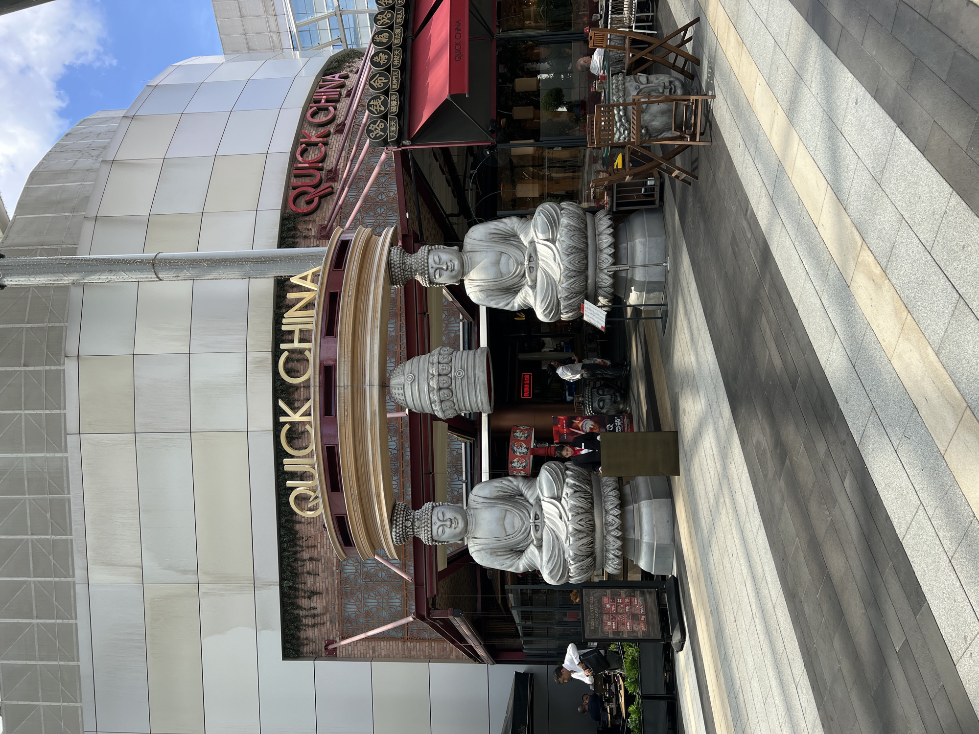This media file captures an outdoor scene in Ataşehir, Turkey. The primary subjects are two large, ornate Buddha statues positioned vertically on pedestals. They are made of a light-colored material and appear to be decorative elements outside a building.
The building facade behind the statues is modern, featuring a white, curved structure with a glass canopy. A red awning with gold lettering that reads "QUICK CHINA" is prominently displayed on the building. The lettering is repeated in gold on a circular structure to the left of the statues.
The ground is paved with large, rectangular tiles in varying shades of grey and beige, laid in a striped pattern. In the background, through the windows of "QUICK CHINA," restaurant seating and tables are visible, along with some potted plants. A few people can be seen inside and near the entrance of the restaurant.
Wooden chairs and tables are placed outside the restaurant, some in front of the statues. The lighting suggests it is daytime, with shadows cast on the paved ground, indicating a sunny day. The scene appears to be a commercial or public space, likely a shopping area or a restaurant district. The overall impression is one of a vibrant, perhaps bustling, urban environment with an Asian-themed establishment.