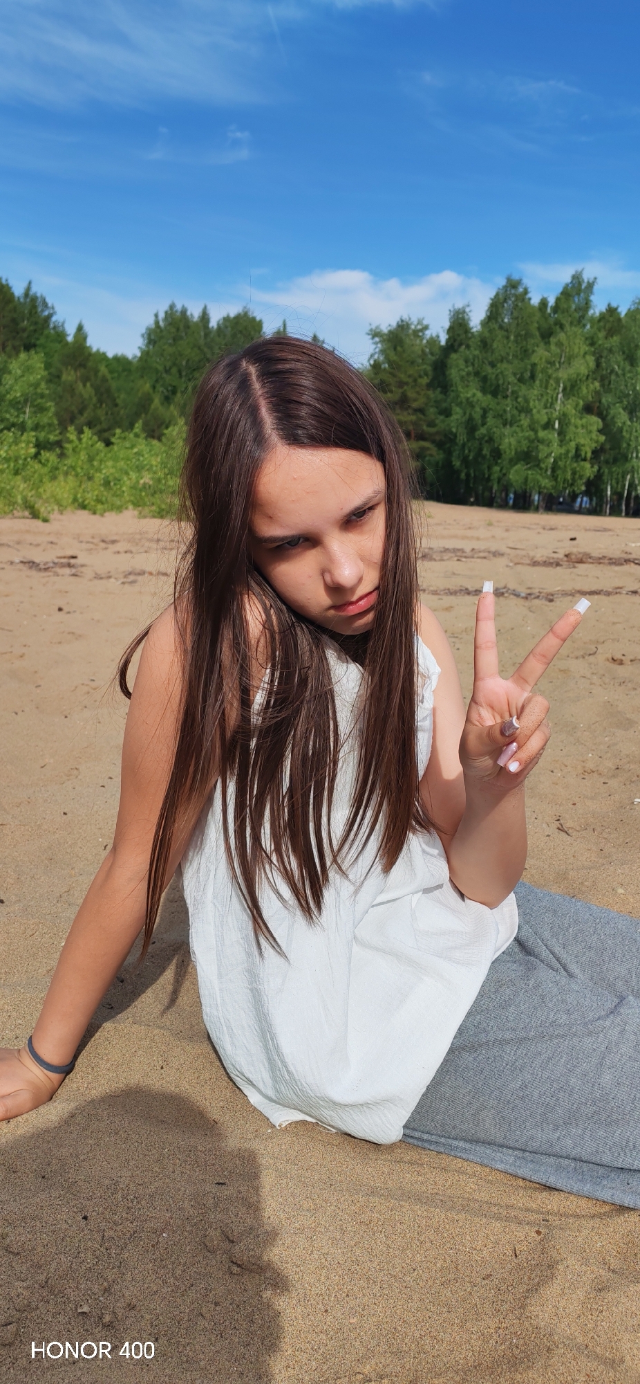 A young girl with long, straight dark brown hair, parted in the middle, is seated on a sandy beach or shore, captured in a brightly lit outdoor portrait. She wears a white sleeveless top and grey pants, and is looking slightly downwards and to her right (viewer's left) with a somewhat serious or "cool" expression. Her right hand is raised, making a 'peace' sign, and her fingernails are painted with a light-colored polish, with some sand visible on the back of her hand. A subtle blue bracelet adorns her left wrist.
The setting behind her is a natural landscape, featuring a line of dense green trees, suggesting a forest bordering the sandy area. Above the treeline, the sky is a clear, vivid blue with only a few very faint, wispy clouds, indicating sunny weather. The strong sunlight casts visible shadows on the sand, consistent with midday or early afternoon. This scene depicts a serene natural environment, characteristic of an outdoor location such as Noviy, Russia. The text "HONOR 400" is visible in the bottom left corner of the image.