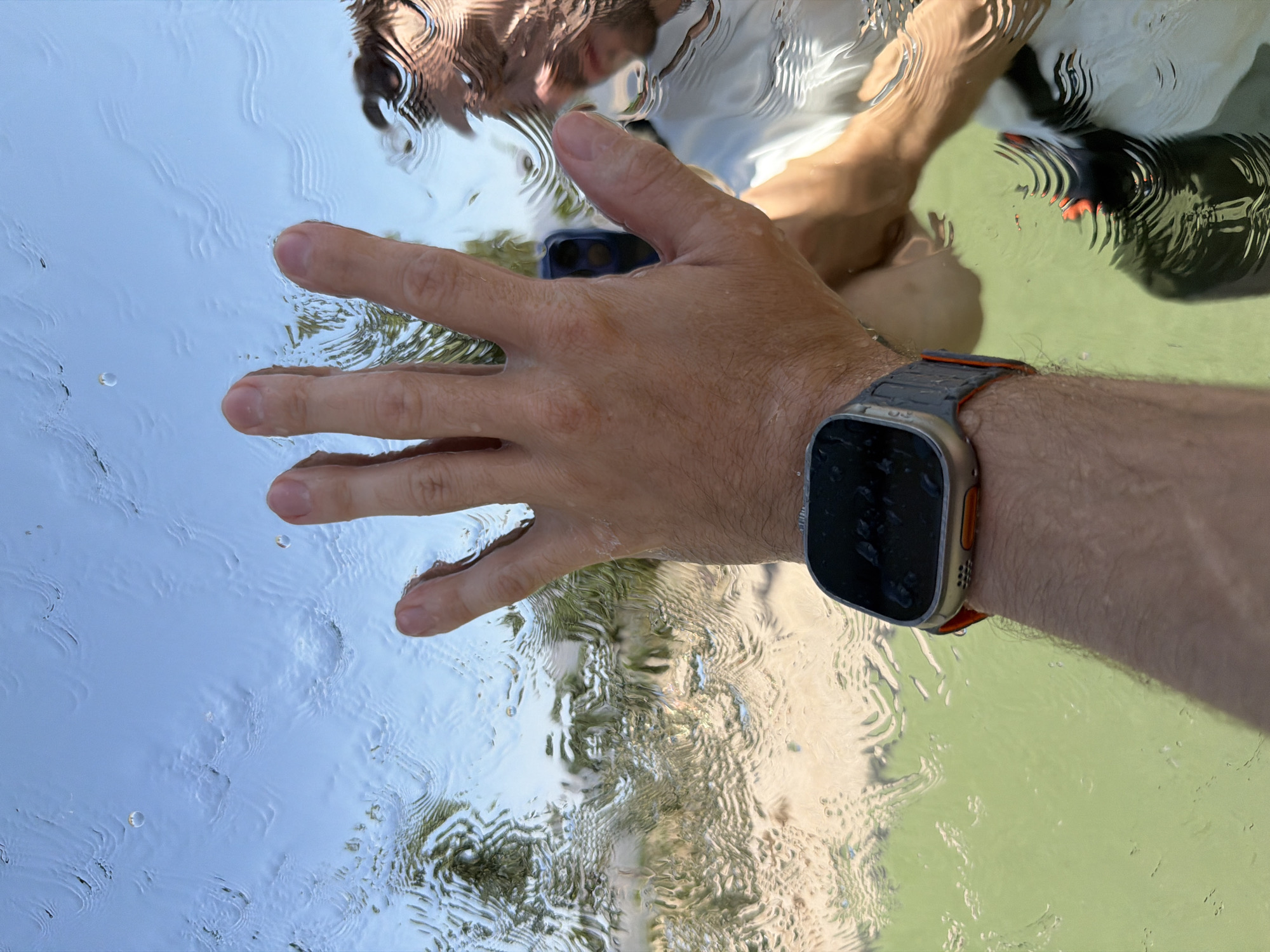 A person's hand is pressed against a wet, reflective surface, possibly a window or a pool cover, distorting the view of the environment beyond. The surface is covered in water droplets and rippling patterns, suggesting it is either raining or submerged in water. The sky is visible, appearing bright blue with some clouds. Below, a green, watery area is visible, possibly a swimming pool, with a hint of sandy ground and foliage. The person is wearing a smartwatch with an orange band on their left wrist. A reflection of the person's face and torso is visible in the wet surface, indicating they are looking through it. The reflection shows them wearing a white shirt. The overall scene has an abstract and somewhat disorienting quality due to the distortion from the water. The context of Charvak, Uzbekistan, is not directly discernible from the visual elements of the image alone; this information would need to be provided externally.