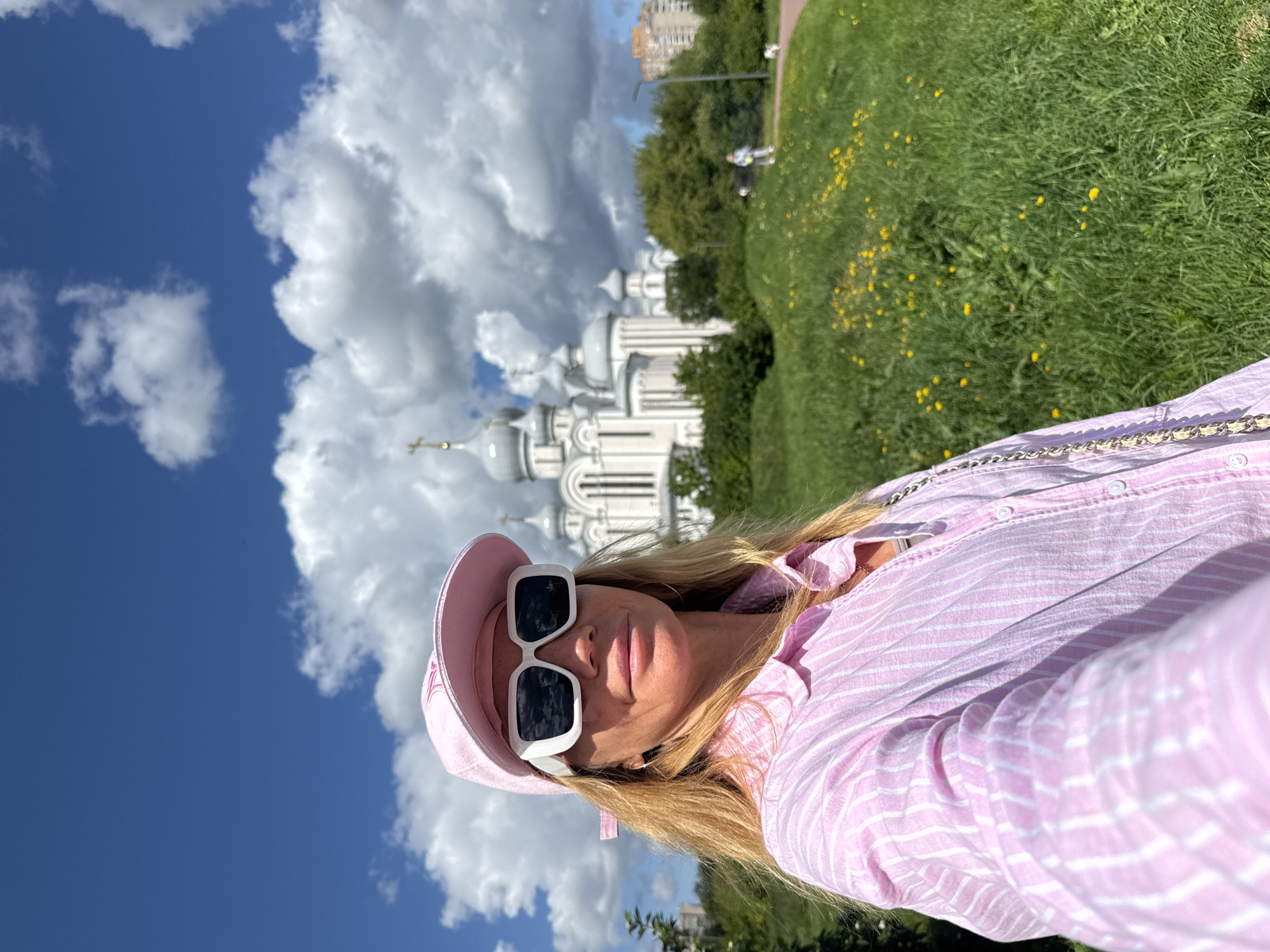 A woman with blonde hair, wearing a pink striped shirt, a pink baseball cap, and large white-framed sunglasses, is taking a selfie outdoors. In the background, a large white building with multiple domes and a cross on top, resembling a church or cathedral, is visible. The building is situated behind a grassy hill dotted with yellow wildflowers. The sky above is bright blue with scattered white clouds. The scene appears to be in an urban park or green space within Saint Petersburg, Russia, suggested by the architectural style of the building and the presence of apartment buildings in the distance. There are other people walking dogs on a path in the middle ground.