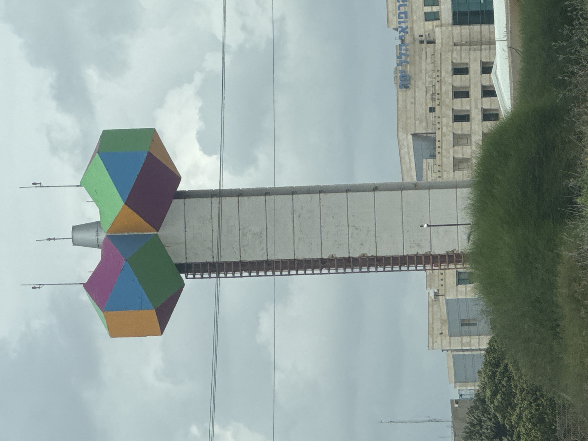 The image depicts a water tower, colloquially known as the "Hadera Testicles," located in Hadera, Israel. The tower is distinguished by two geodesic domes painted in various colors, situated atop a concrete shaft. A walkway with a metal railing extends from the shaft. A building with Hebrew text visible is adjacent to the tower. Overcast skies are present in the background. Vegetation is seen to the right of the tower. Electrical wires are visible running across the scene.