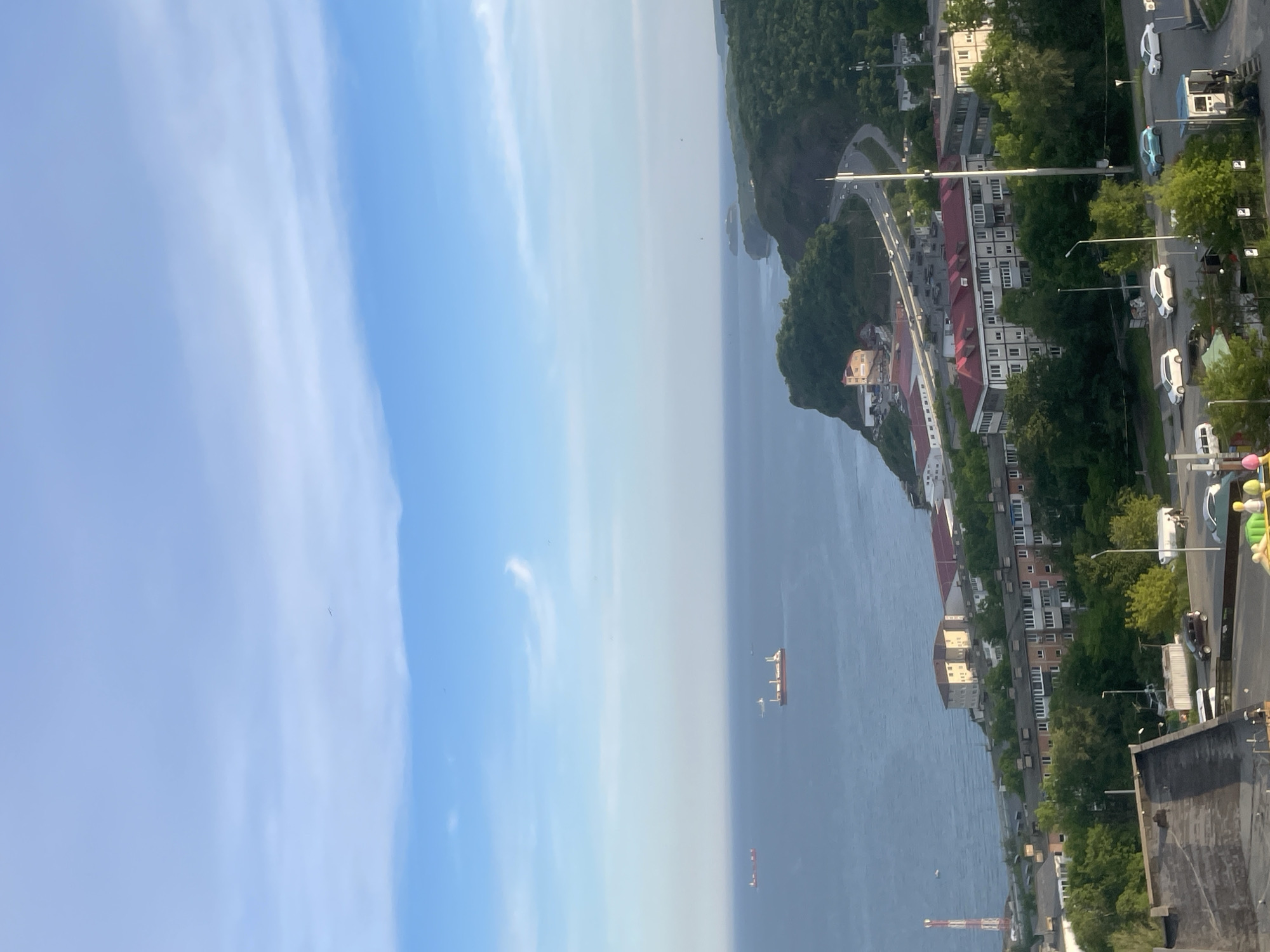 This is an aerial shot of a coastal city, likely Vladivostok, Russia, on a clear day. In the foreground, a cluster of multi-story residential buildings with red roofs is visible, interspersed with trees and a road with several cars. A tall, red and white striped tower stands to the left of these buildings. Beyond the buildings, a body of water stretches to the horizon. Several cargo ships are present on the water. The coastline is characterized by rolling, green hills, with a winding road visible on one of them. The sky is blue with scattered white clouds.