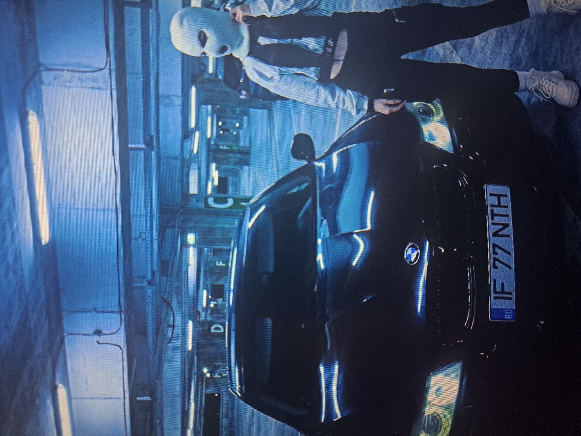 The image depicts a person wearing a ski mask leaning against the hood of a dark-colored BMW car. The car has a Romanian license plate that reads "IF 77 NTH". The background suggests an indoor parking garage, illuminated by fluorescent lights. The environment is stark and functional, consisting of concrete walls and ceilings with visible piping and signage. There are no identifiable features to indicate the location in Yerevan, Armenia. The focus is on the car and the masked individual.