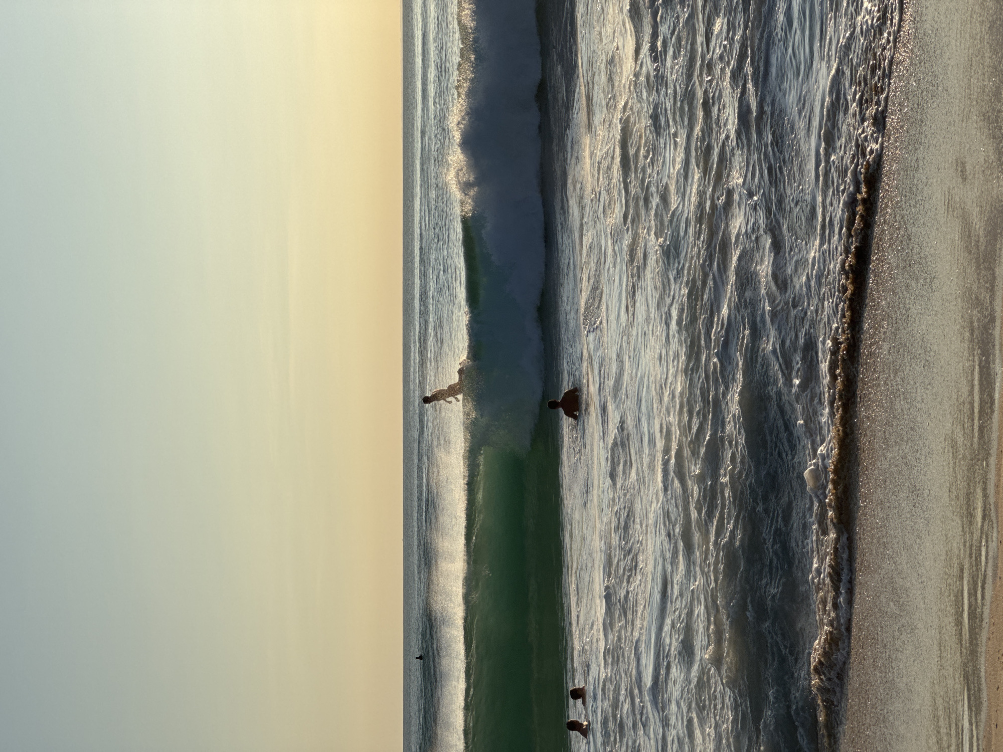 This is a vertical beachside view along the shoreline of Kuta Selatan, Indonesia. The sky is a gradient of light blue to pale yellow, likely indicating sunset or sunrise. Three people are visible in the water. One person is riding a wave, and the others are either swimming or standing in the wave. The ocean is choppy with multiple waves heading towards the shore. The beach is sandy, and the edge of the water is marked by a line of darker sand.